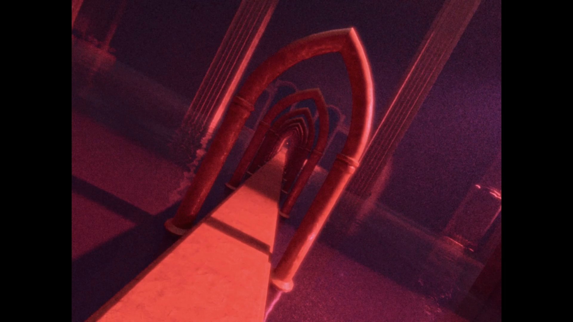 An ethereal camera movement traverses a series of repeating gothic arches and stone pillars bathed in deep red light. The sequence creates a rhythmic, hypnotic progression through a dark, monumental corridor.