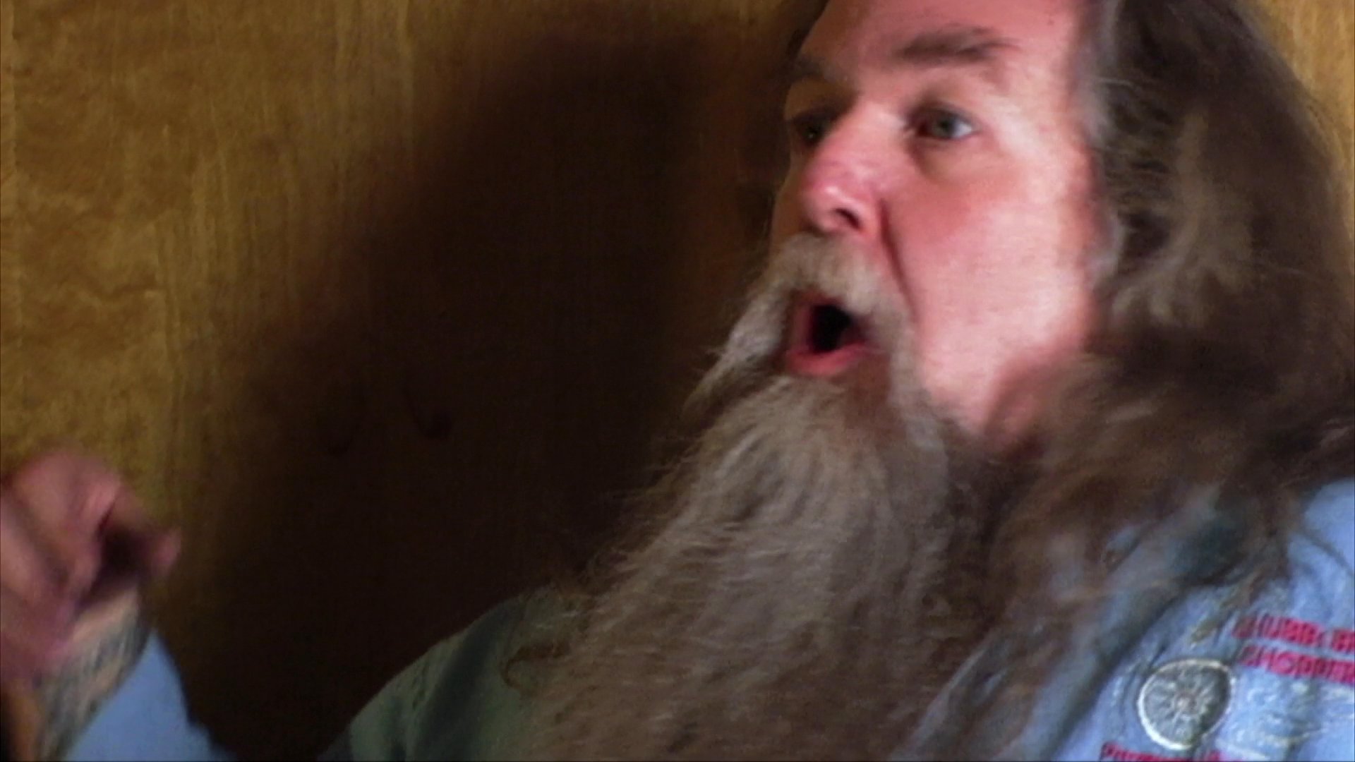 A man with a long beard uses animated gestures and varied facial expressions while narrating a story. He maintains engagement through dynamic physical communication against a simple wooden background.