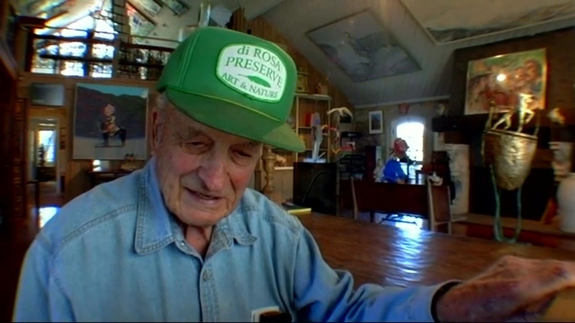 An elderly man in an art-filled room recalls becoming more interested in visual art than writing after arriving on the Left Bank. He presents the green woman portrait as the first picture he ever bought.