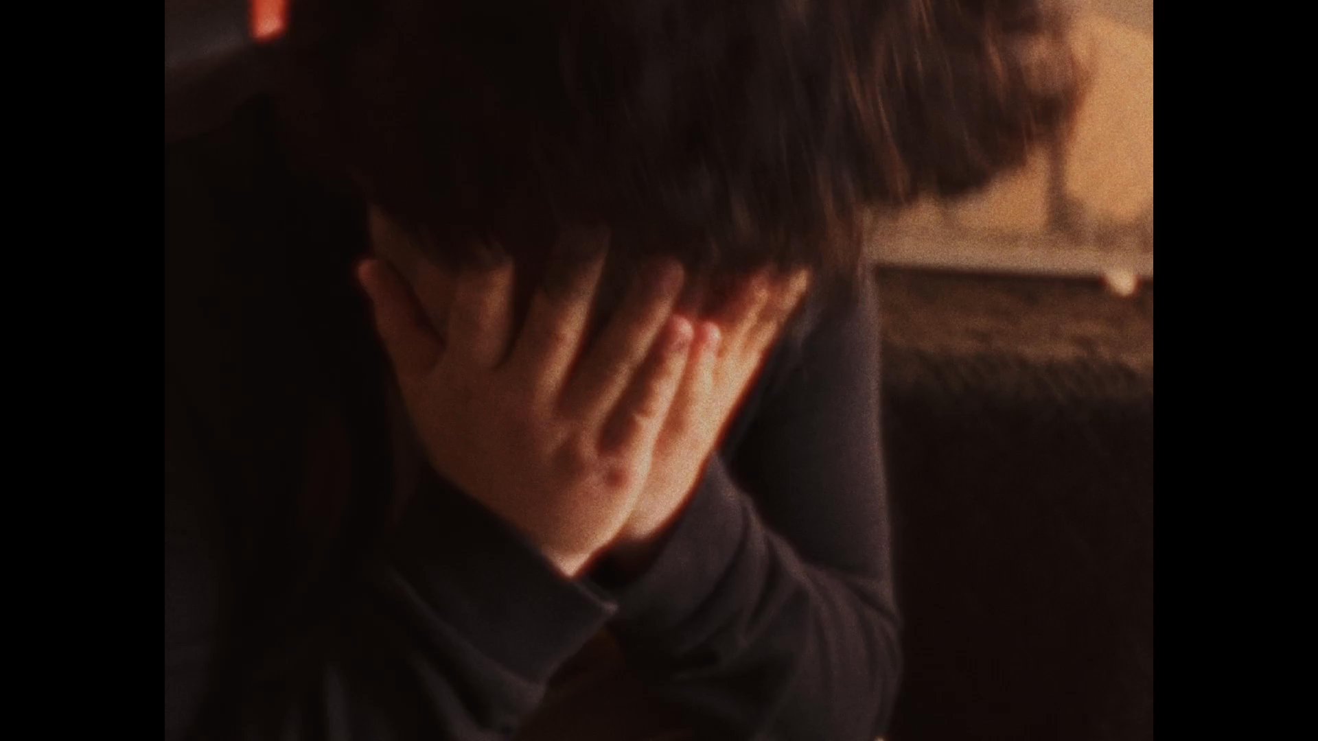A young man sits alone in his room, appearing visibly distressed and covering his face with his hands. The scene shifts to an aerial perspective of a house, emphasizing a theme of isolation.