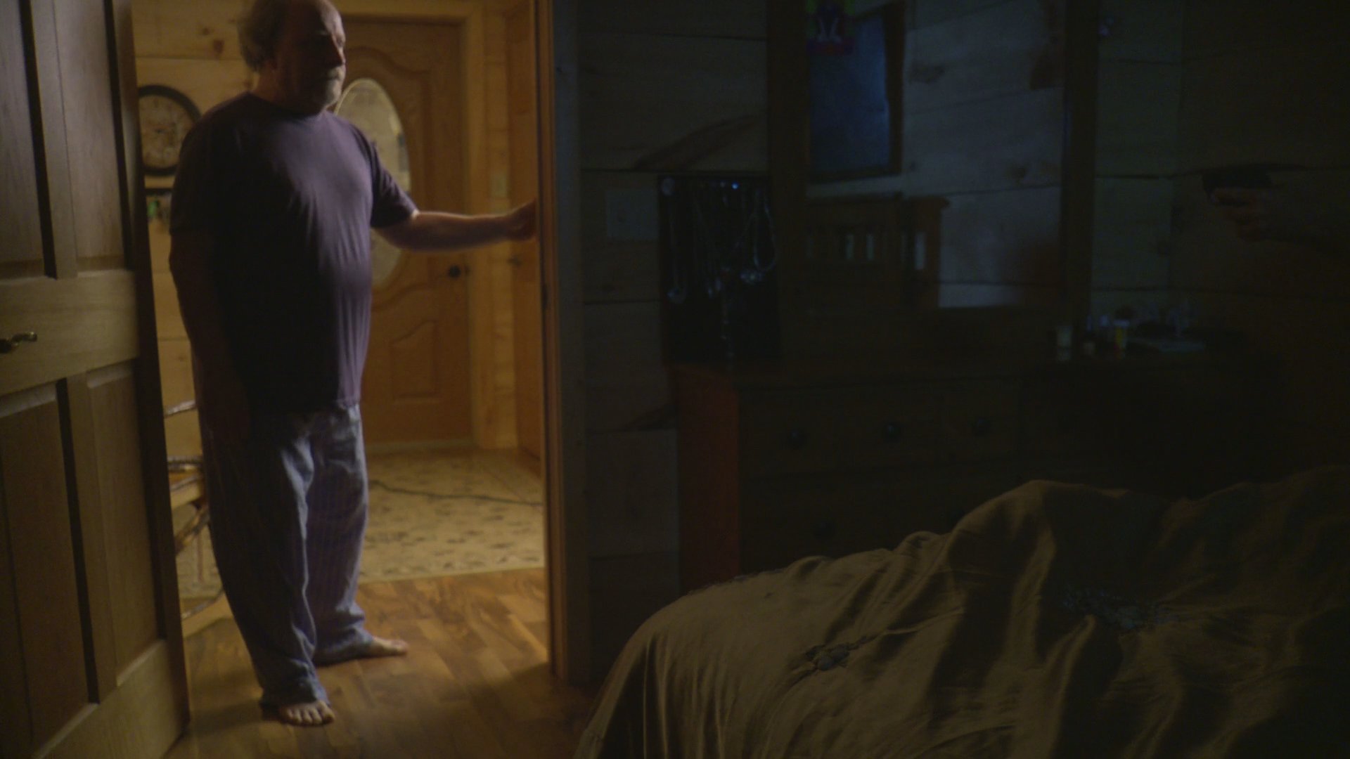 A woman in a bedroom aims a handgun at a man entering the room, resulting in a brief struggle before she maintains her defensive position.