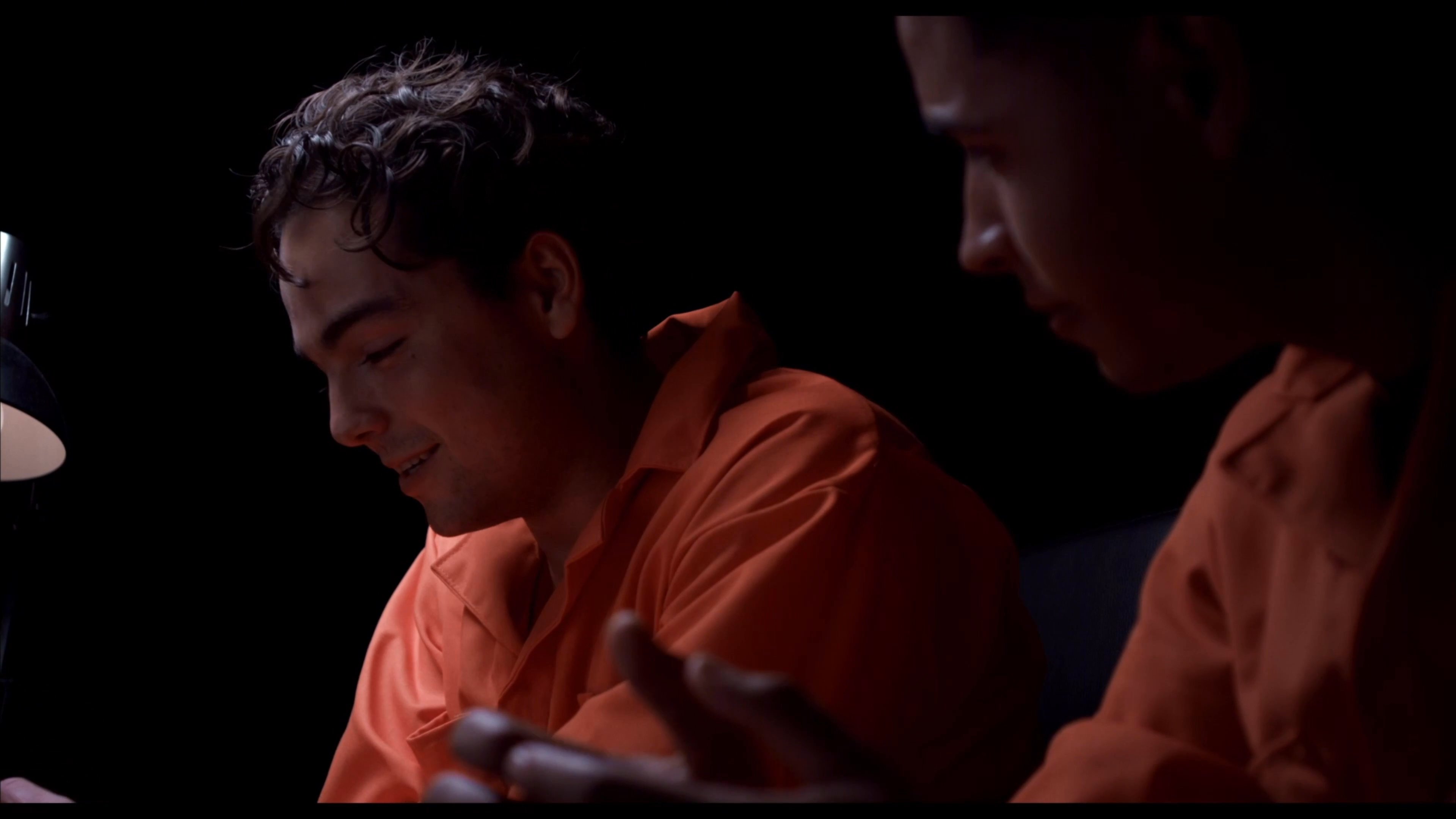 Two individuals wearing orange jumpsuits engage in a serious, low-light conversation within a confined, dark setting. The interaction emphasizes intense interpersonal communication and emotional gravity.