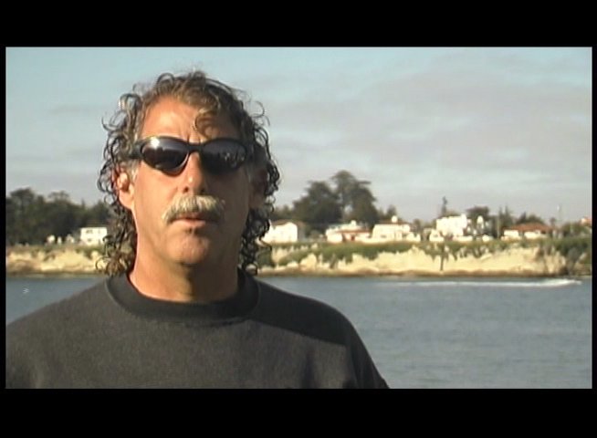 A man wearing sunglasses and a mustache speaks to the camera against a coastal backdrop, followed by a shot of a surfboards and wetsuits shop.