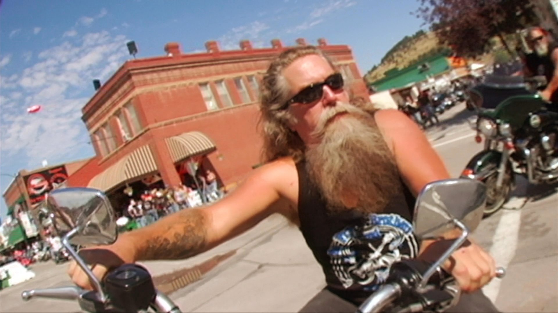 A man performs with an acoustic guitar before transitioning to footage of a long-haired, bearded motorcyclist riding through a crowded rally in a town center.