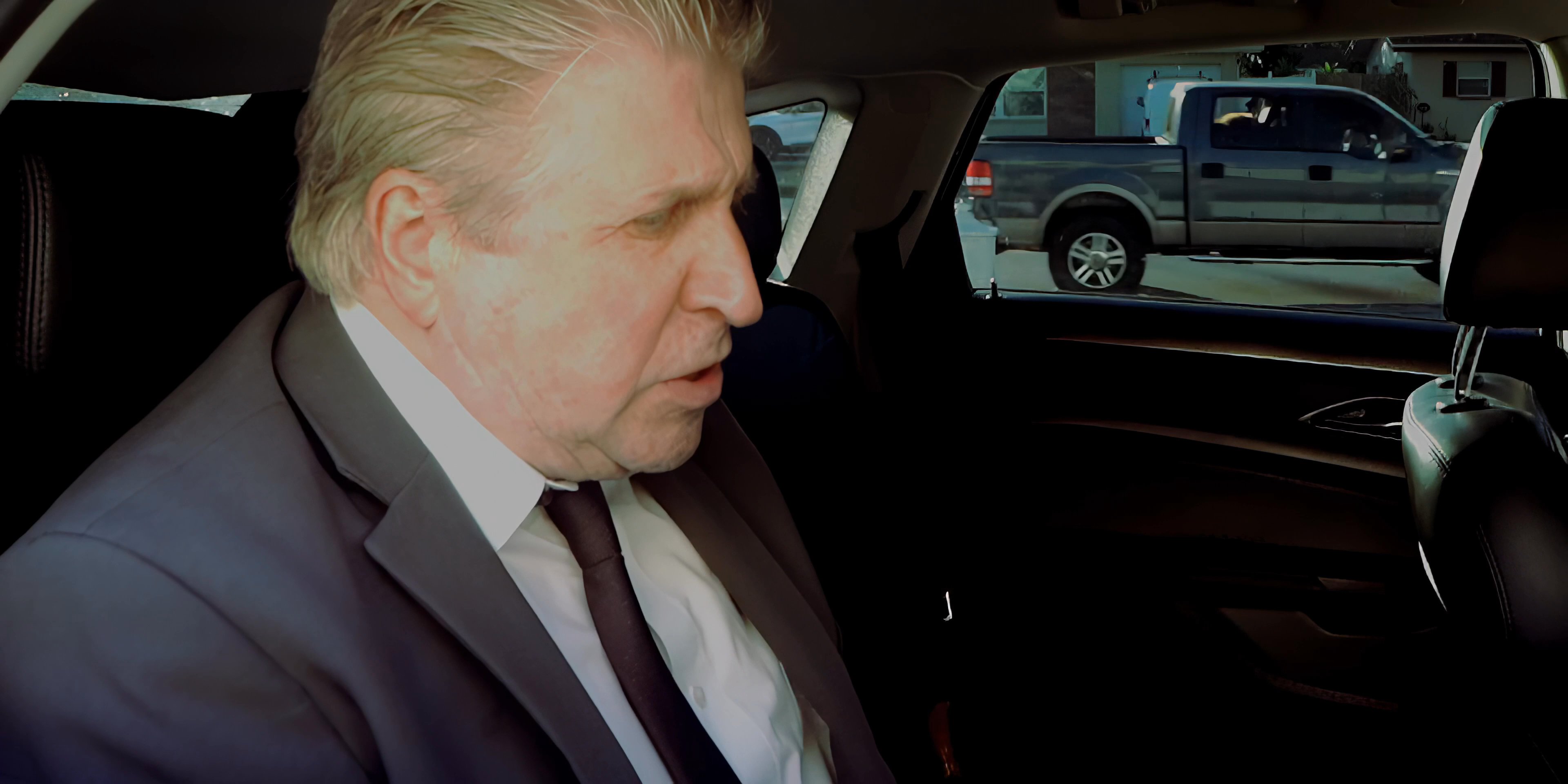 An older man in a suit drinks water in the backseat while conversing with the driver of the vehicle. The scene highlights a professional or formal interaction occurring during a commute.