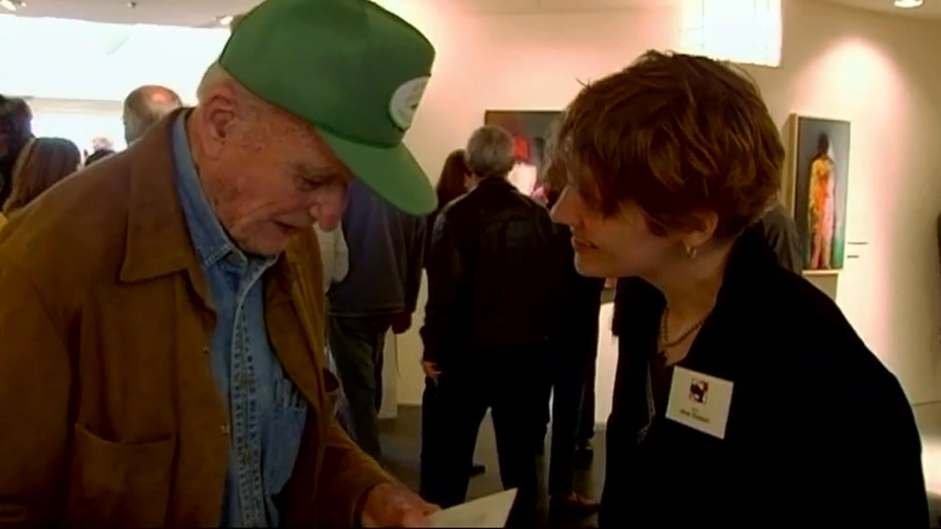 At a crowded art gallery, an older visitor speaks with artist Jenna Gress and discusses her piece “Shiver.” He offers $400 for the artwork while a short-haired gallery attendant listens nearby.