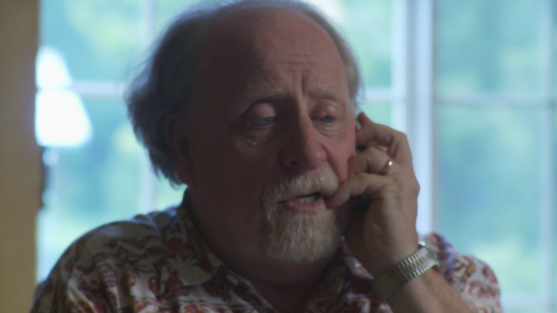 An elderly man exhibits visible signs of emotional distress while engaged in a somber, serious telephone conversation near a window.