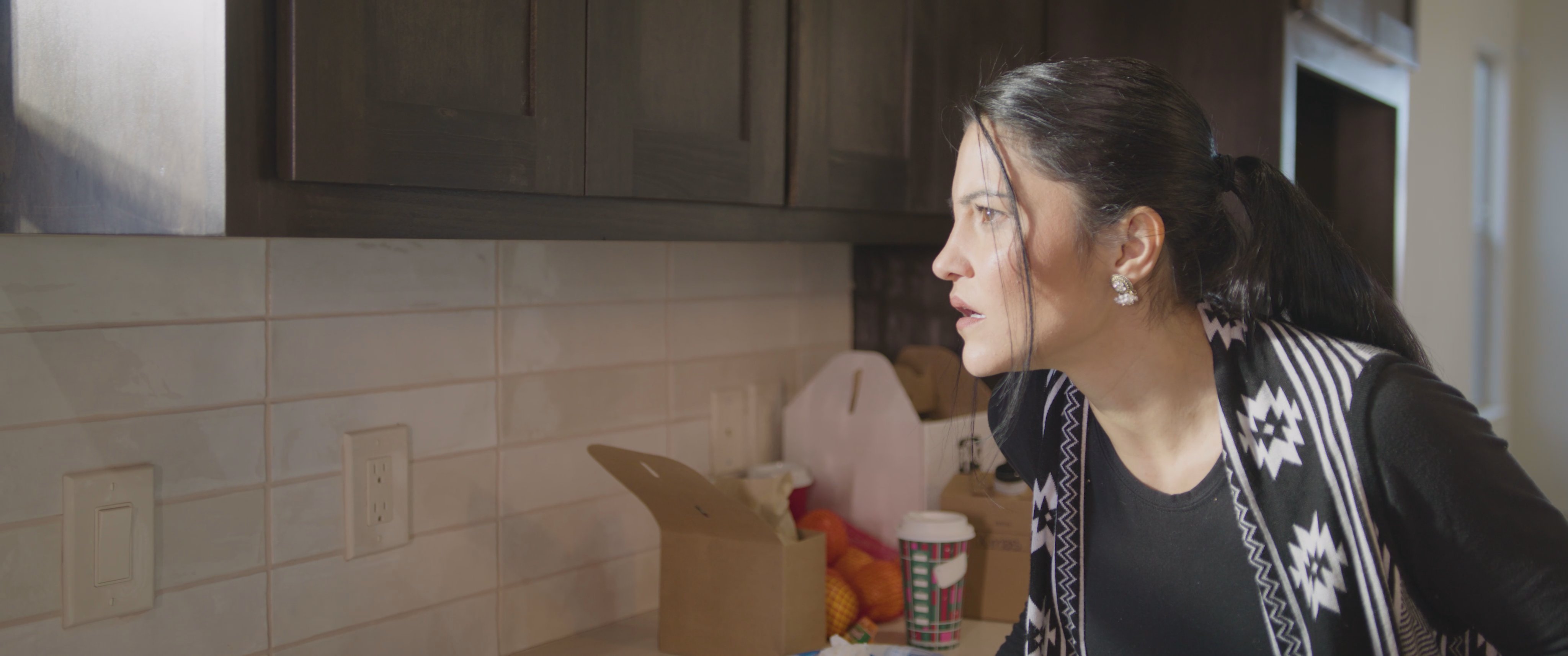 A woman looks out from her kitchen window to monitor a construction worker outside before returning to her morning routine. She observes the outdoor activity with focused attention and unease.