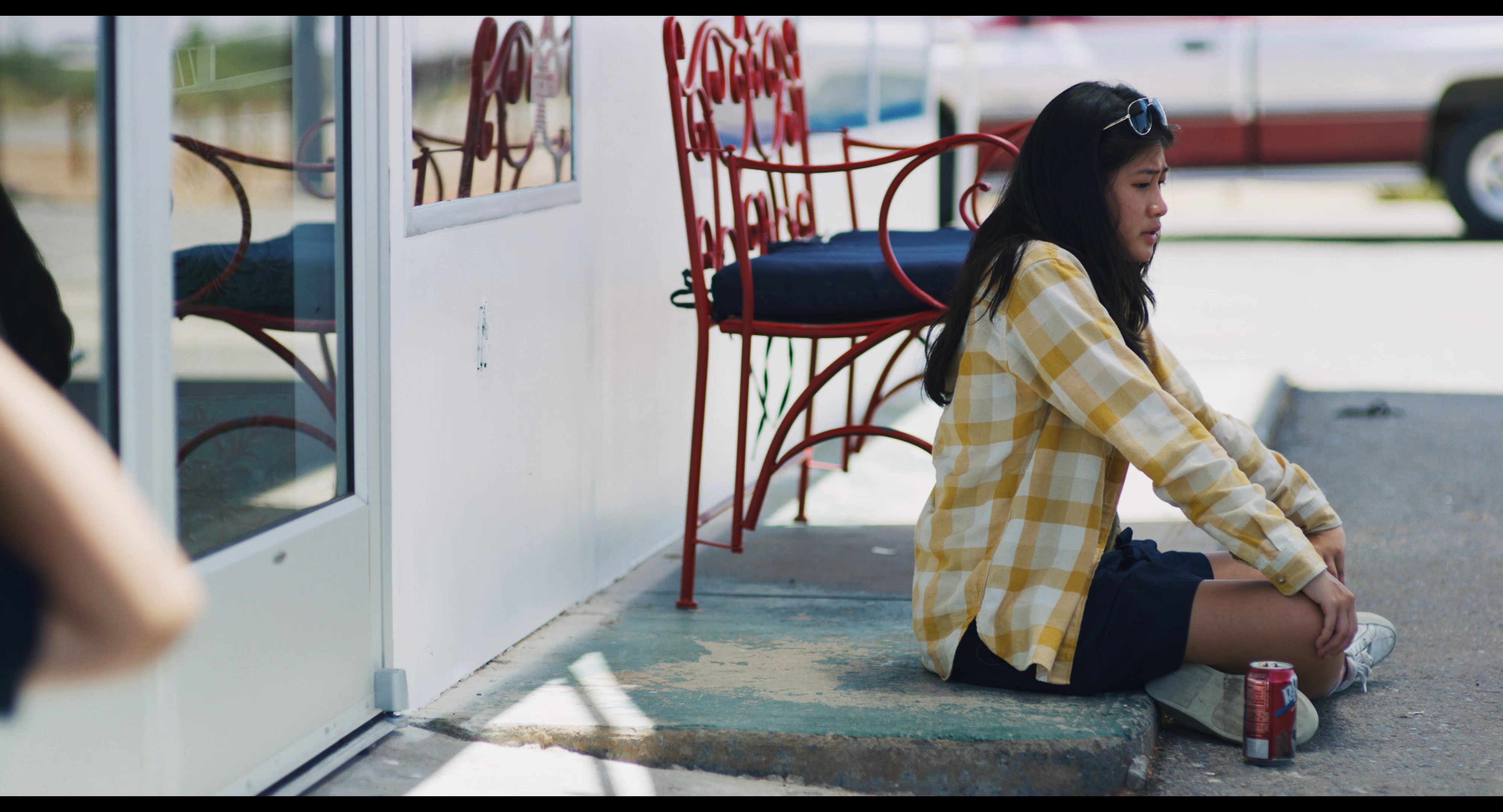A young woman sits on the pavement outside a laundromat entrance while a man waits nearby. The scene captures a quiet moment of anticipation or reflection in an urban setting.