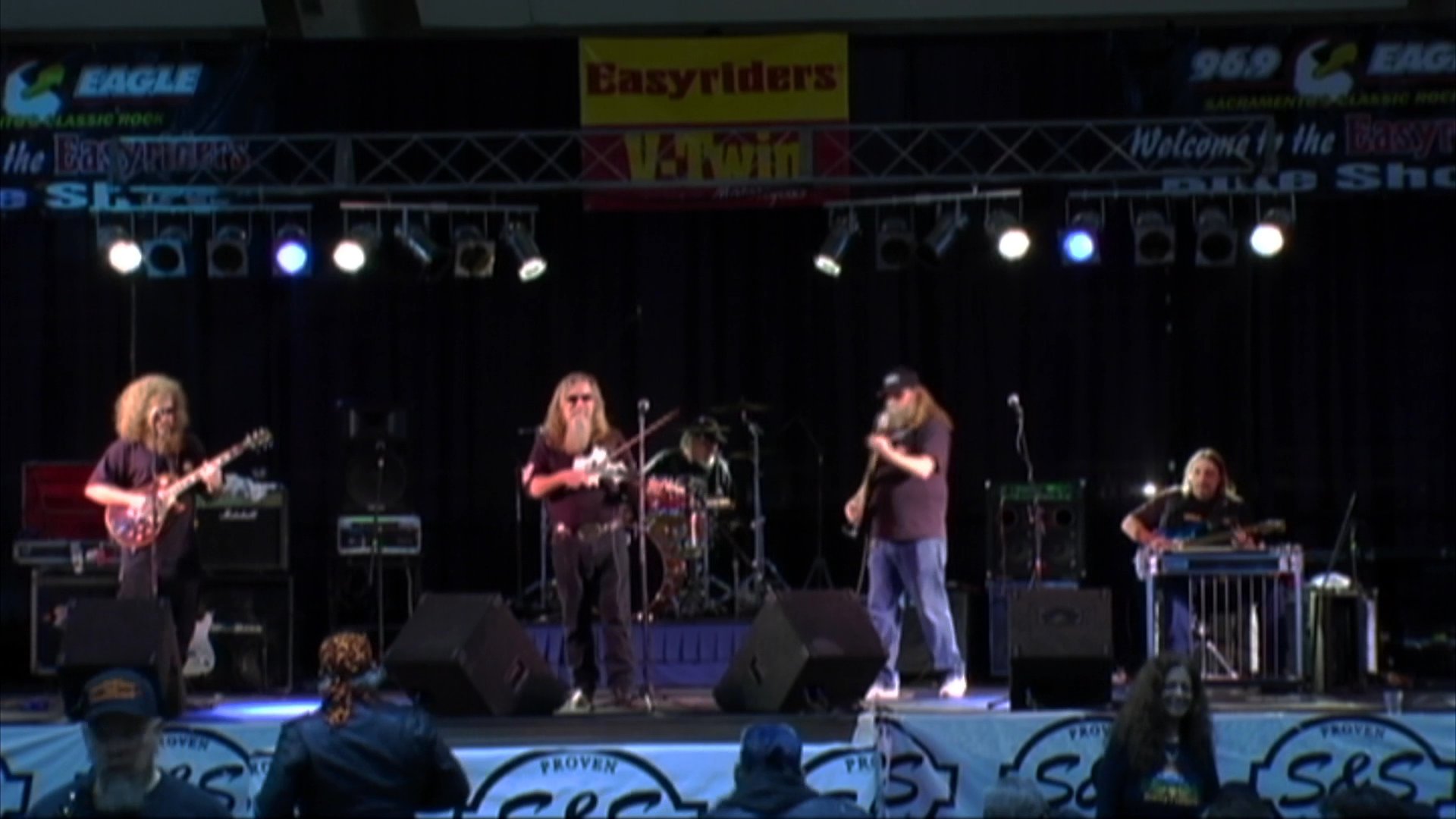 A country band performs on a concert stage, featuring close-ups of musicians playing the pedal steel guitar, fiddle, and electric guitar. The group showcases their instrumental skills in a live setting with professional sound equipment.