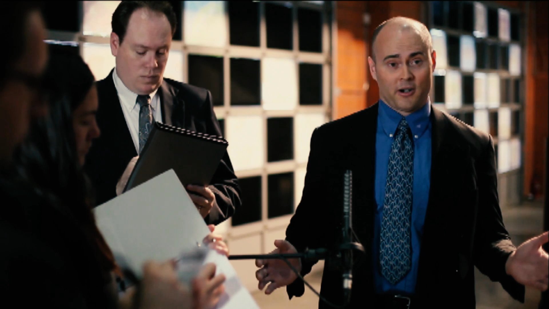 A formal spokesperson in a business suit addresses a group of reporters during an indoor press engagement. He answers questions and provides statements while standing in front of a patterned backdrop.