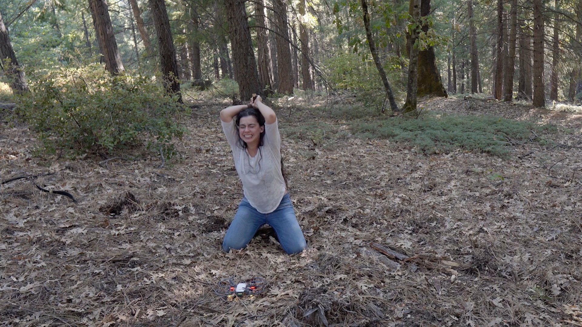 A woman kneeling on the forest floor displays intense emotional anguish and physical distress. She raises her hands in frustration before collapsing into a state of grief.