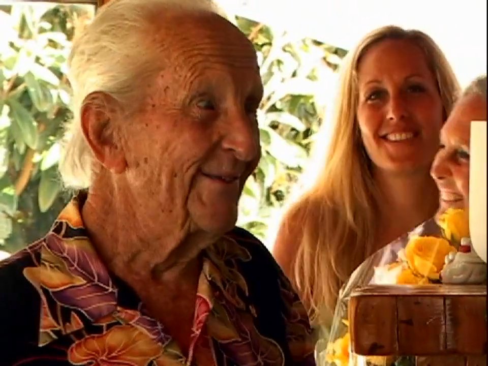 An elderly man receives a heartfelt homecoming surprise from visiting family members. The group exchanges emotional greetings and embraces in a warm, domestic setting.