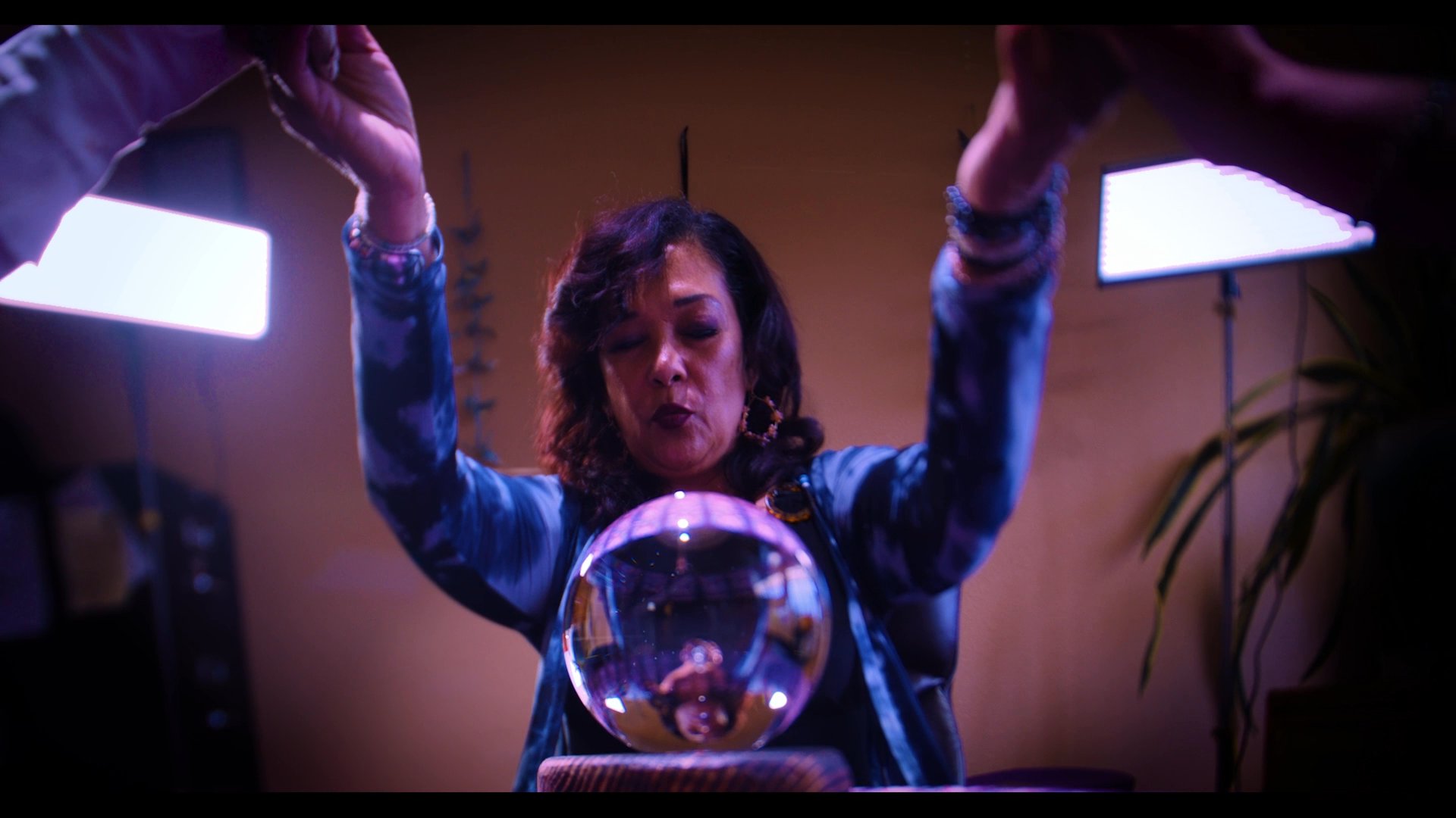 A medium performs a mystical ritual with a client, focusing on a crystal ball to gain insights. The atmosphere shifts to a more grounded setting as a man observes the proceedings.