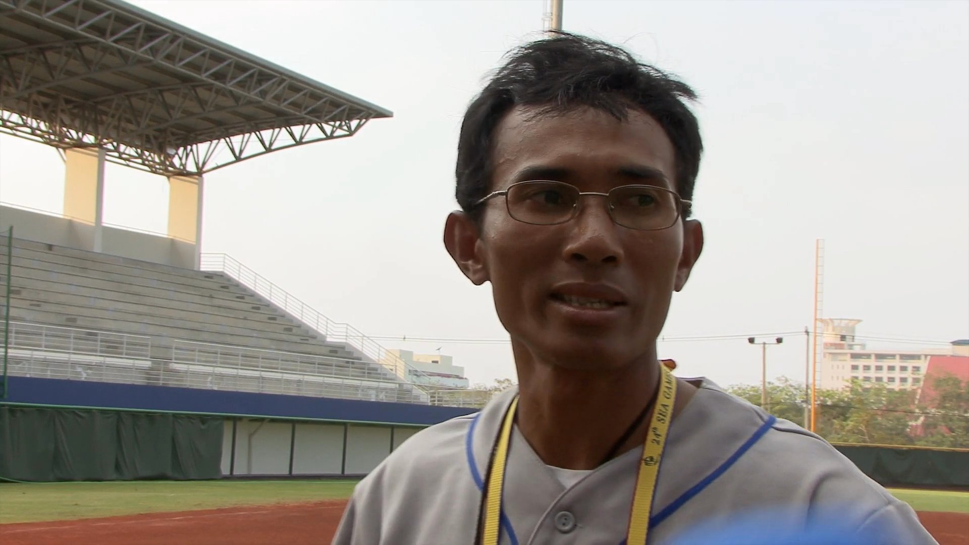 A baseball coach provides commentary while standing on a professional field, using hand gestures to describe gameplay or strategy.