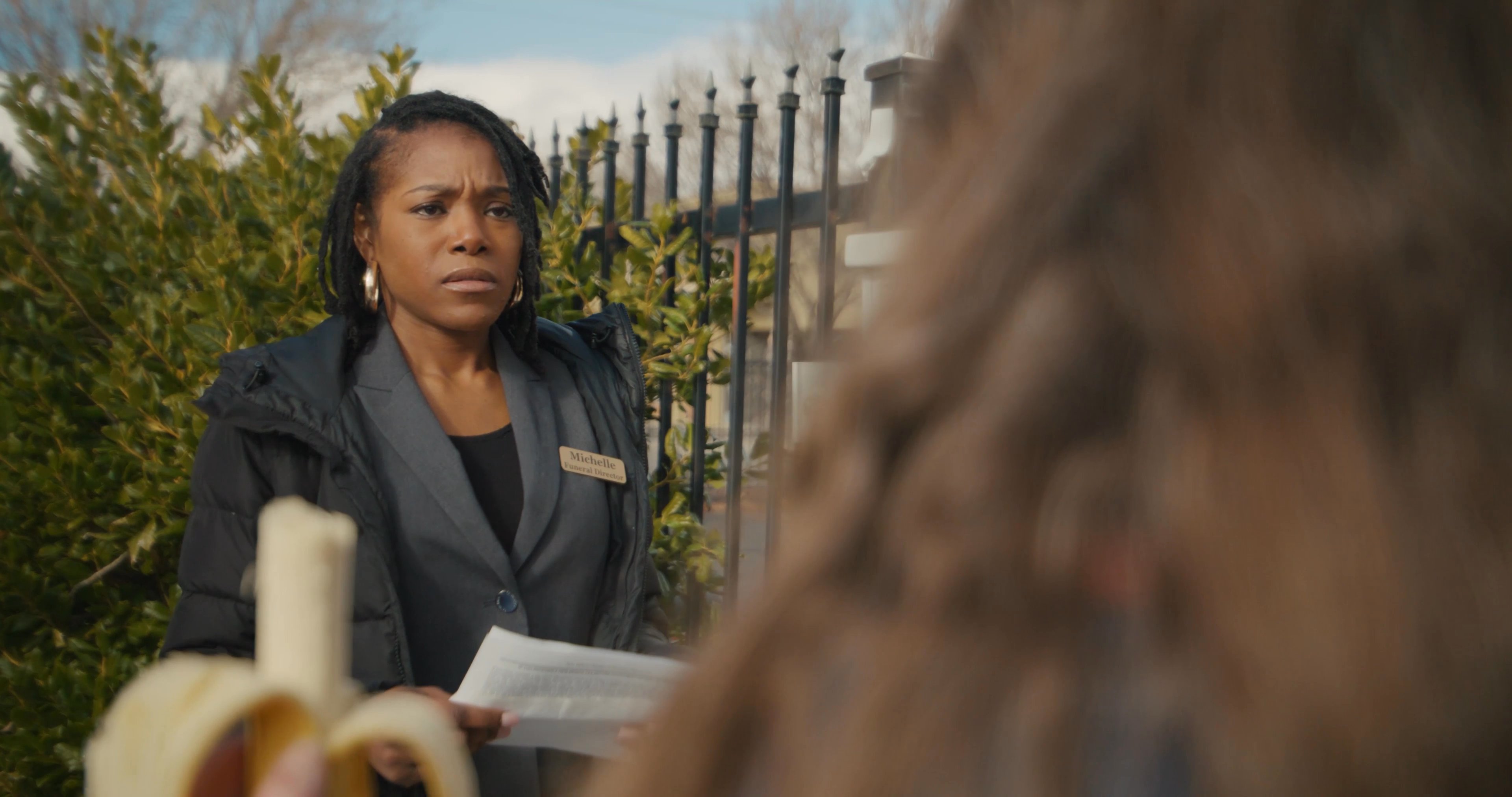 A woman holding a prescription pill bottle and a banana engages in a tense conversation with a professional individual on a city street.