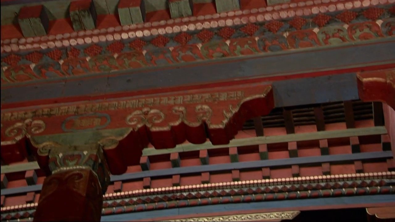 Individuals walk through the courtyard of a traditional Tibetan monastery set against a backdrop of mountainous terrain. The scene highlights intricate wooden architectural details and the serene, elevated environment of the region.