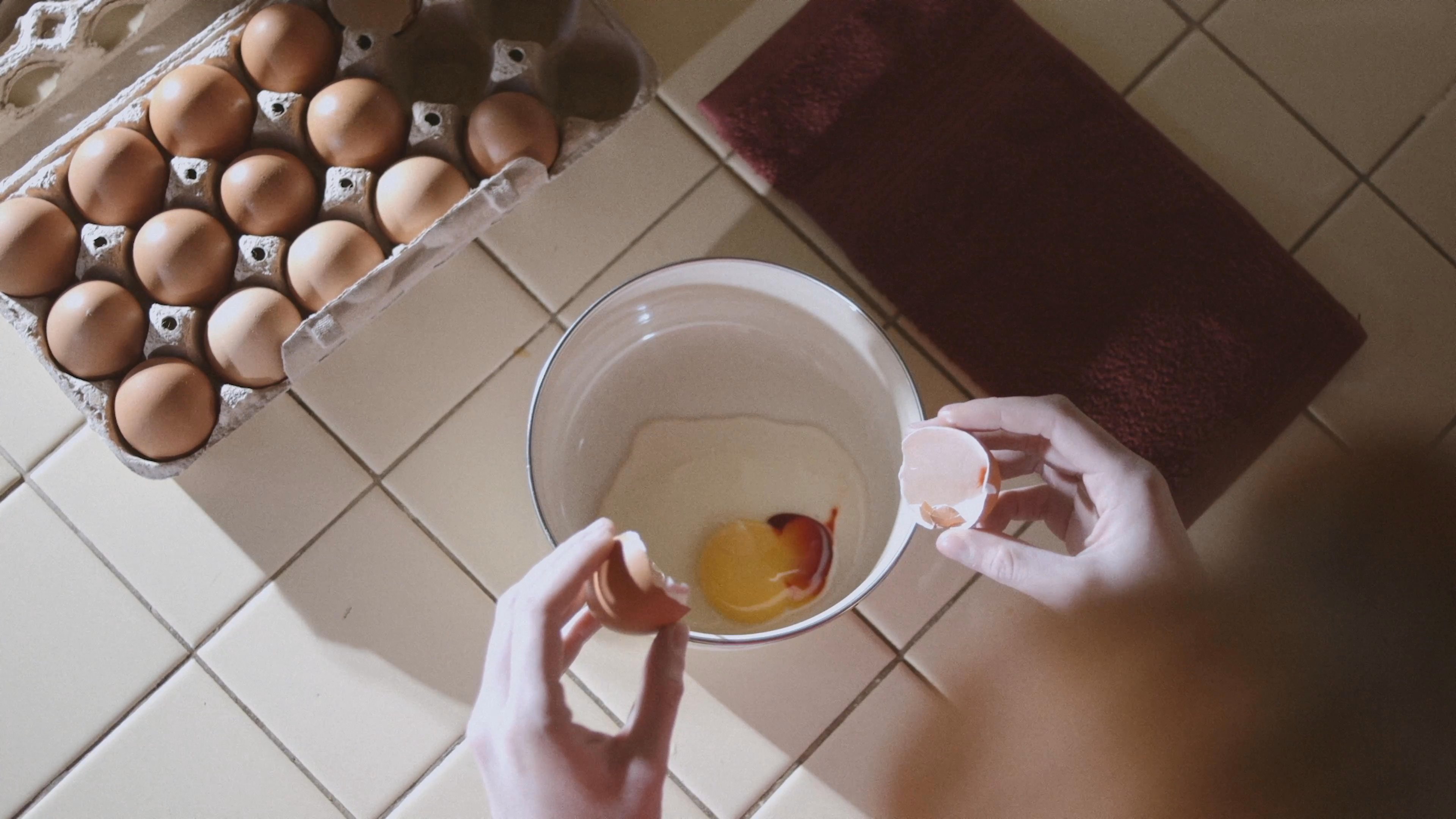 A woman with red hair prepares food in a kitchen, methodically cracking multiple eggs into a mixing bowl.