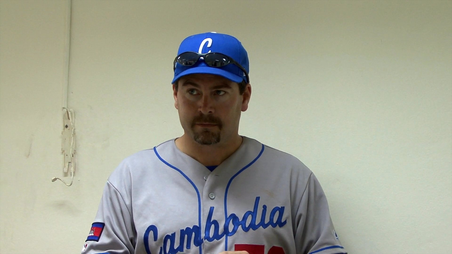 A coach provides tactical instruction to members of the Cambodian national baseball team in a locker room setting. The players listen attentively as the staff outlines strategies for their upcoming performance.