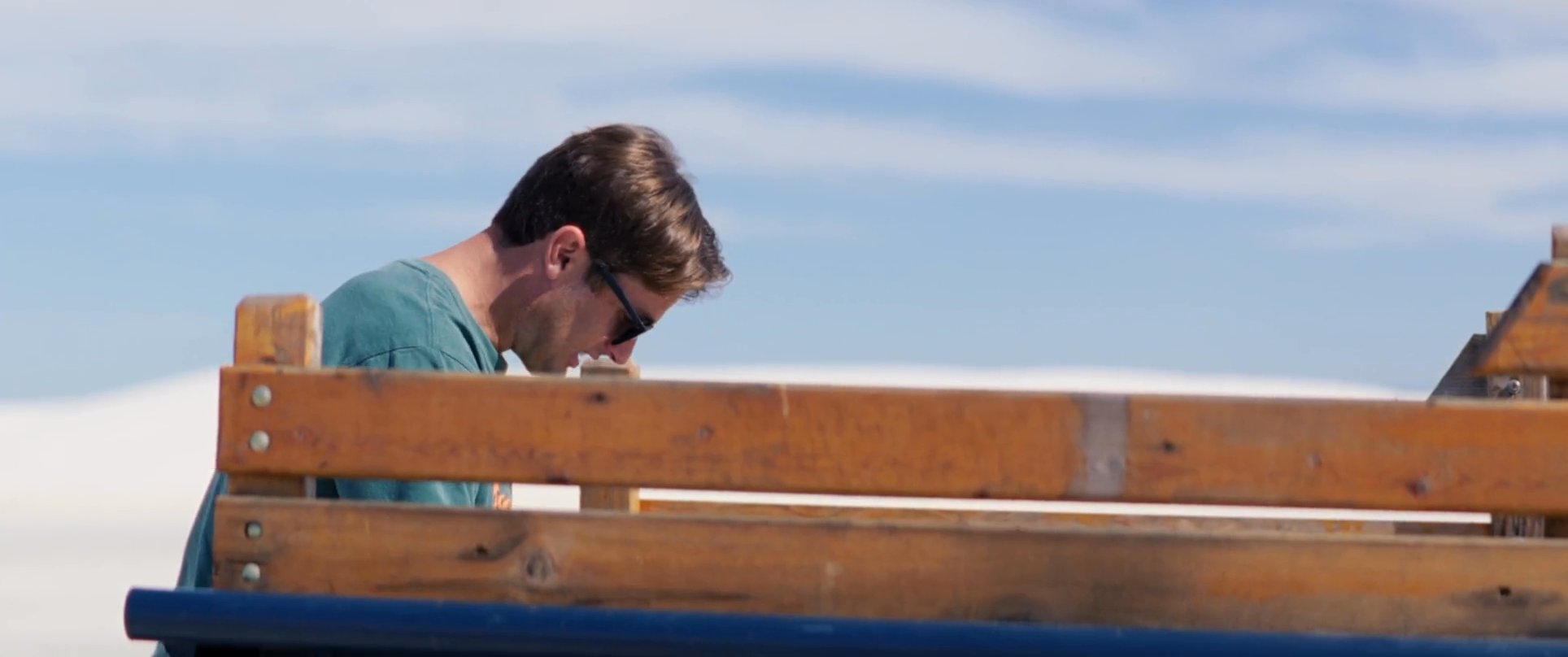 Two men interact while gathered around a vintage pickup truck in an open, desert-like landscape. The scene captures a lighthearted exchange as they unload cargo from the truck bed.