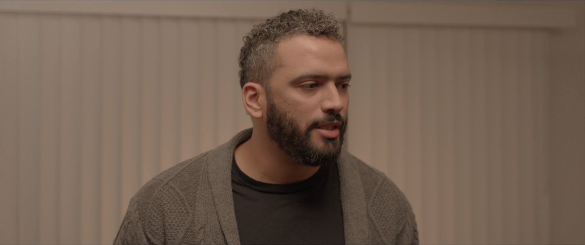 A man engages in a focused conversation, alternating between speaking and listening to a group of men in an indoor setting. The scene highlights the dynamics of an interpersonal exchange.