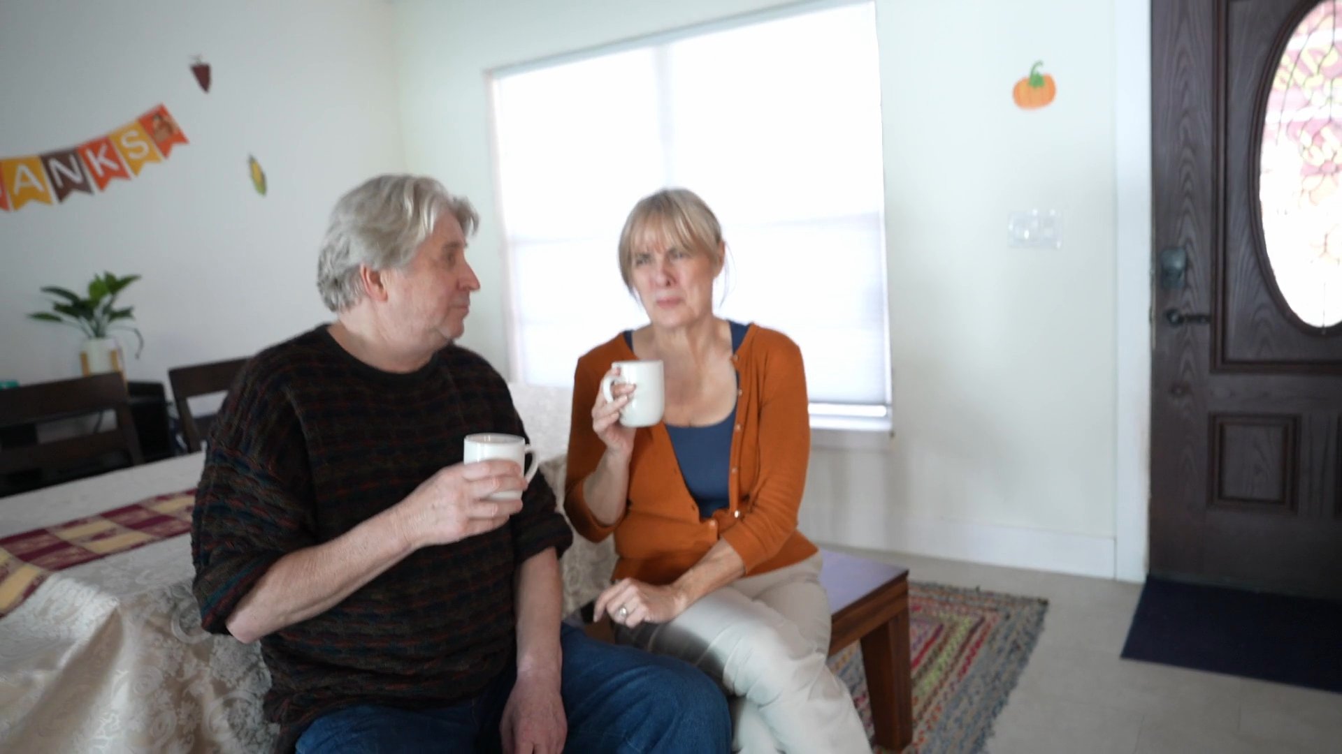 A woman walks through her home, interacts with family members holding coffee mugs in the living room, and proceeds to prepare a drink in the kitchen.