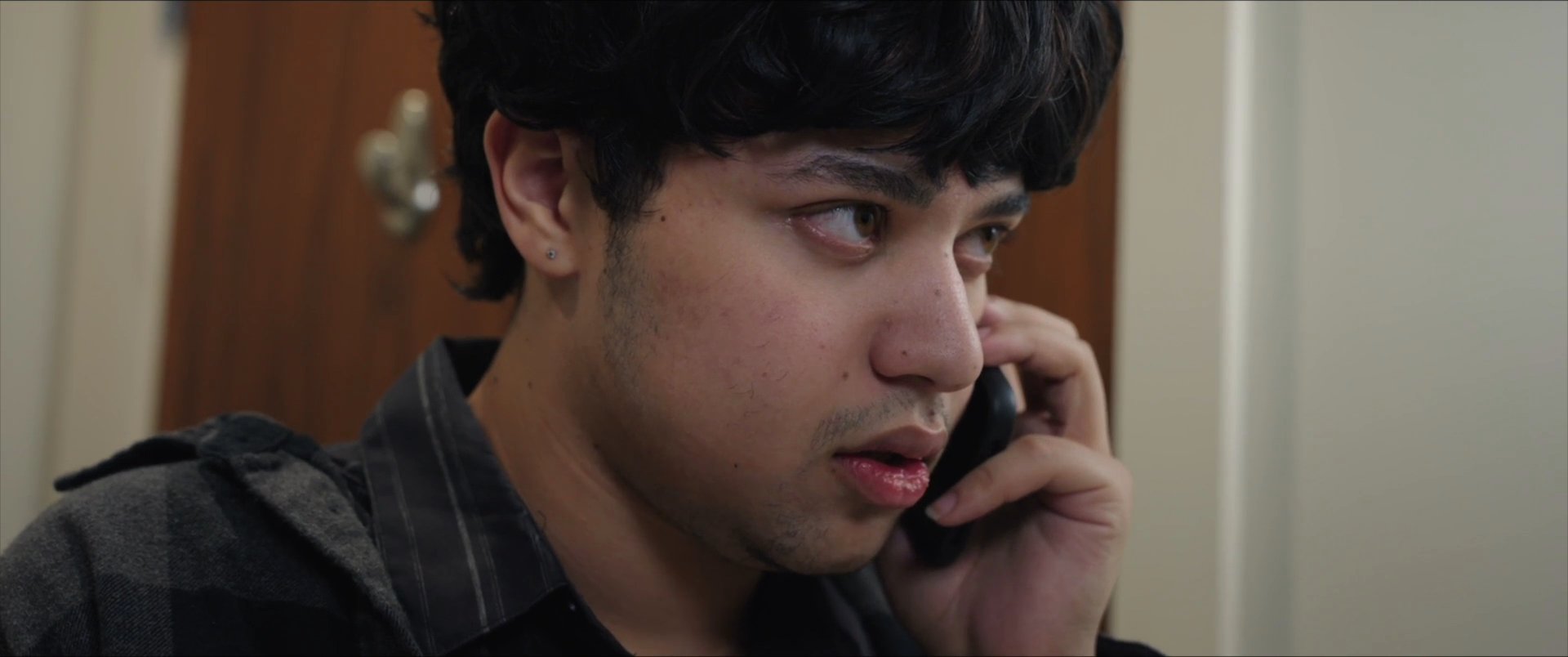 A young man maintains a focused, serious expression while participating in an engaged telephone conversation indoors.