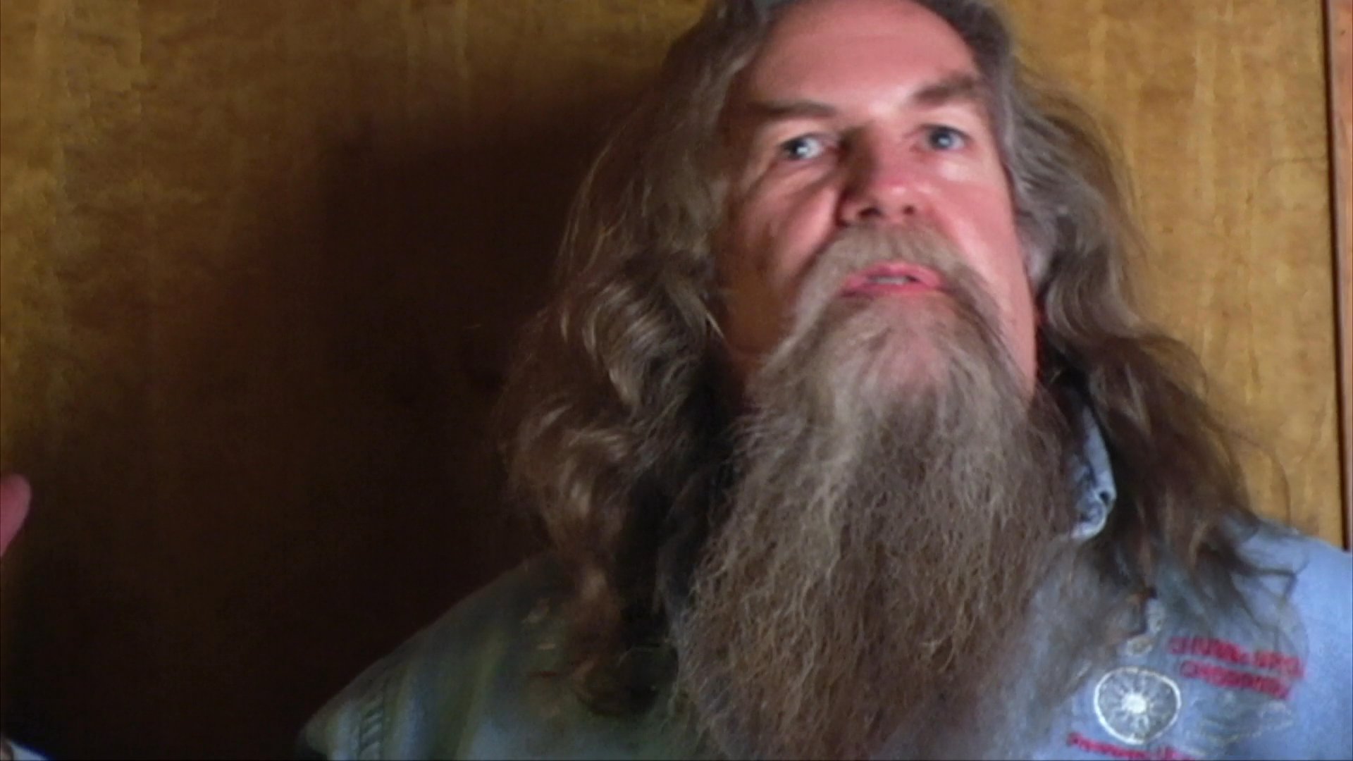A long-haired, bearded man sits against a wood-paneled wall, using expressive hand gestures to recount experiences and demonstrate musical techniques.