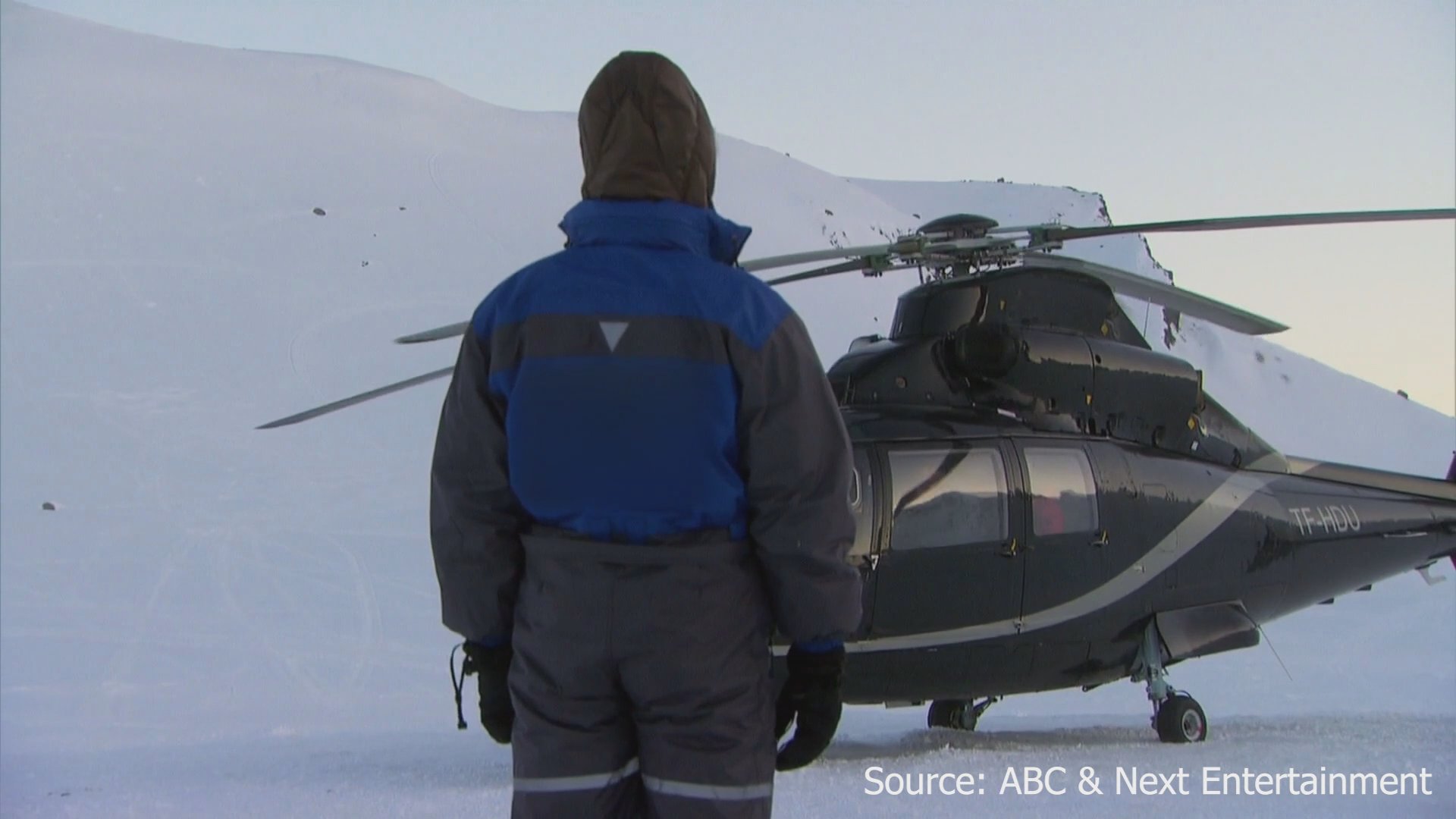Participants in winter gear prepare for departure as a helicopter sits in a snowy, desolate landscape. The group boards the aircraft to travel across the frozen terrain.
