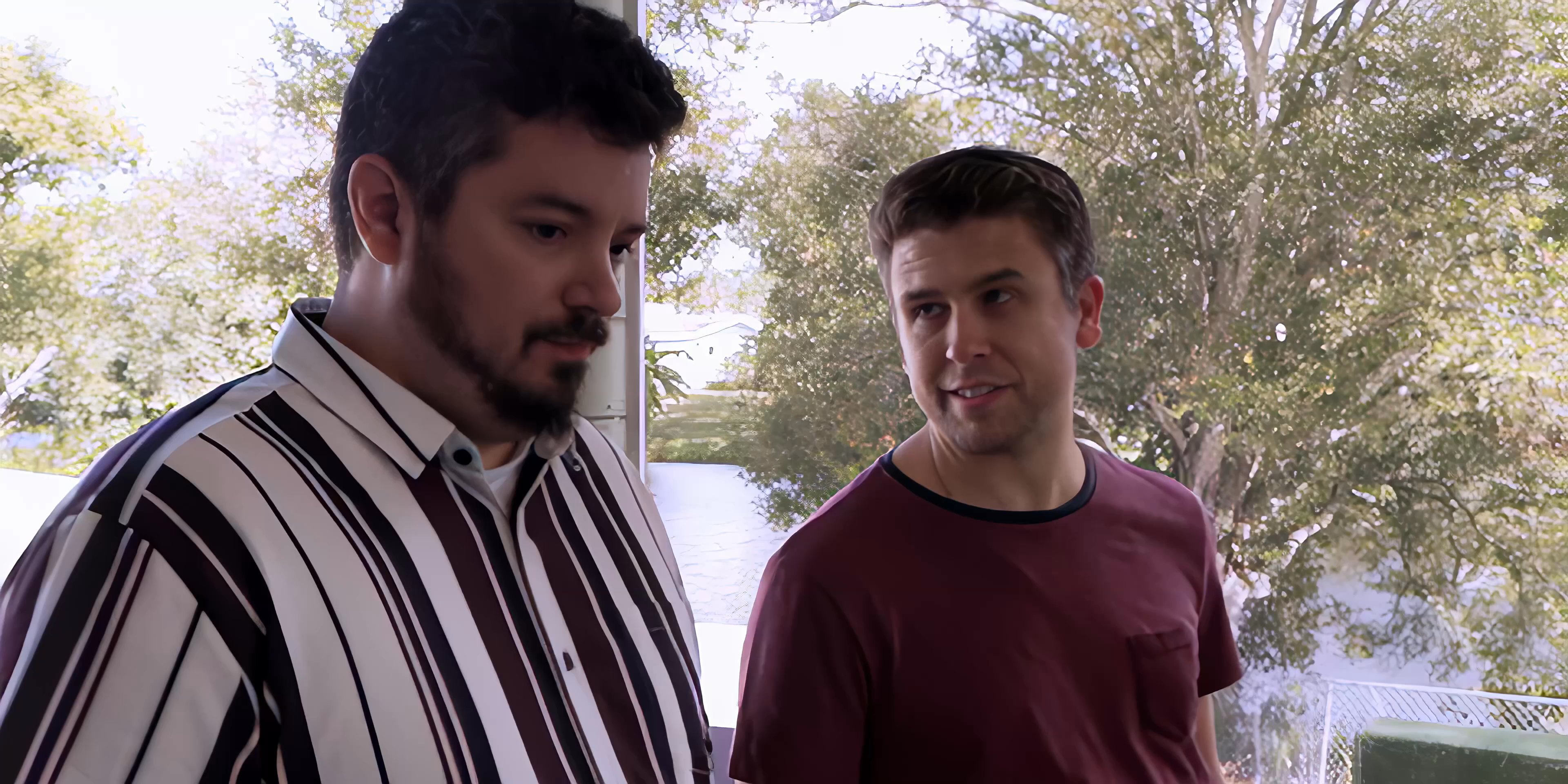 Two men engage in an earnest, focused conversation while standing before a large window overlooking trees.