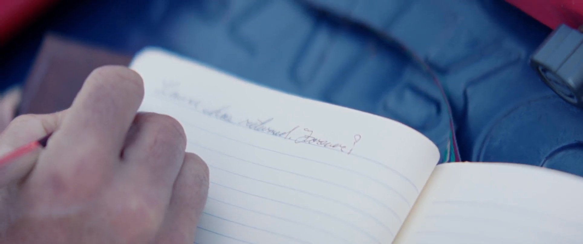 An individual records thoughts in a leather-bound journal while preparing for a camping trip in a scenic, coastal environment. His companion waits nearby as he organizes his gear and finishes his entry.