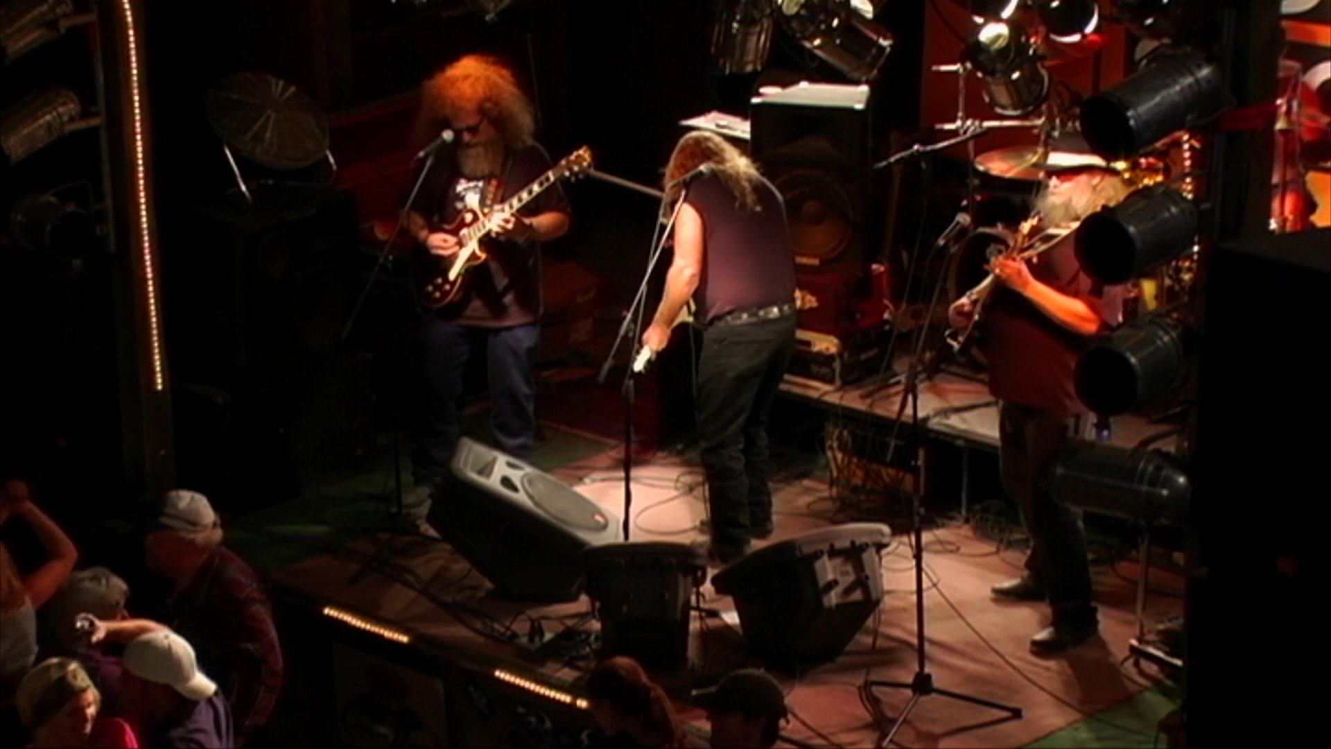 A band performs live music on a stage for an audience in a dim, atmospheric music venue. Musicians play guitars and other instruments while a lead vocalist sings into a microphone.