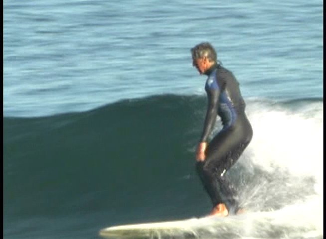 An experienced surfer in a wetsuit maneuvers across a breaking ocean wave. The footage concludes with a brief portrait of the surfer standing on shore.