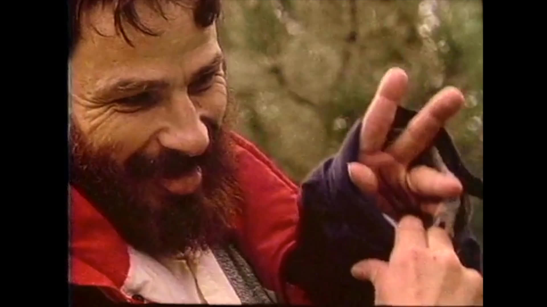 A bearded man performs detailed hand gestures and sign language, focusing intently on his movements while outdoors.