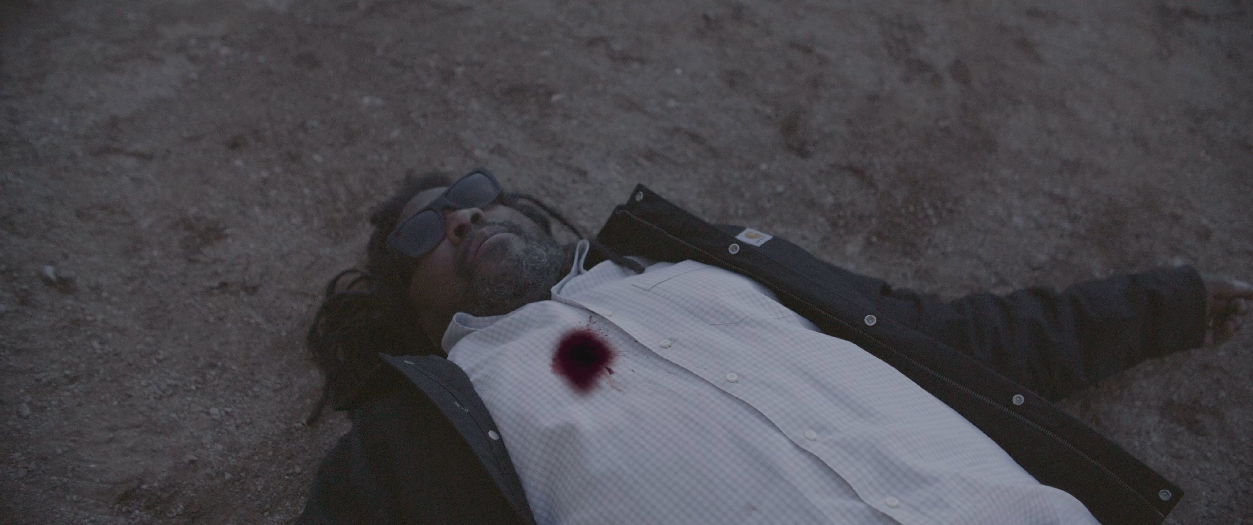 A distressed woman discovers a body in a desert setting and reacts with intense emotional anguish. The scene progresses to reveal a crime-related struggle and a burial site.