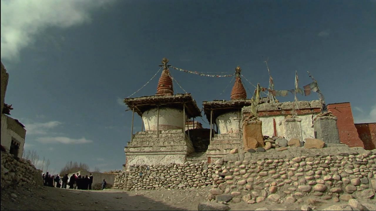 King Jigme Palbar Bista speaks in an interview, set against a backdrop of traditional Himalayan architecture and prayer flags fluttering in a mountain landscape.