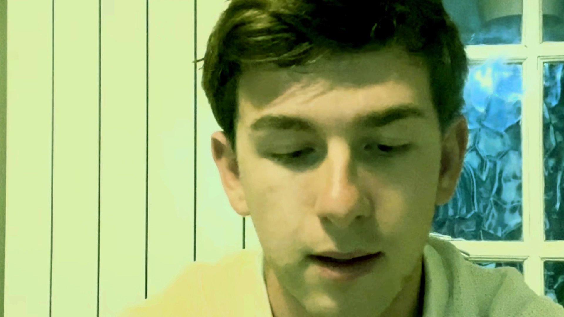 A young man delivers a direct address, maintaining steady eye contact while speaking in a casual indoor setting.
