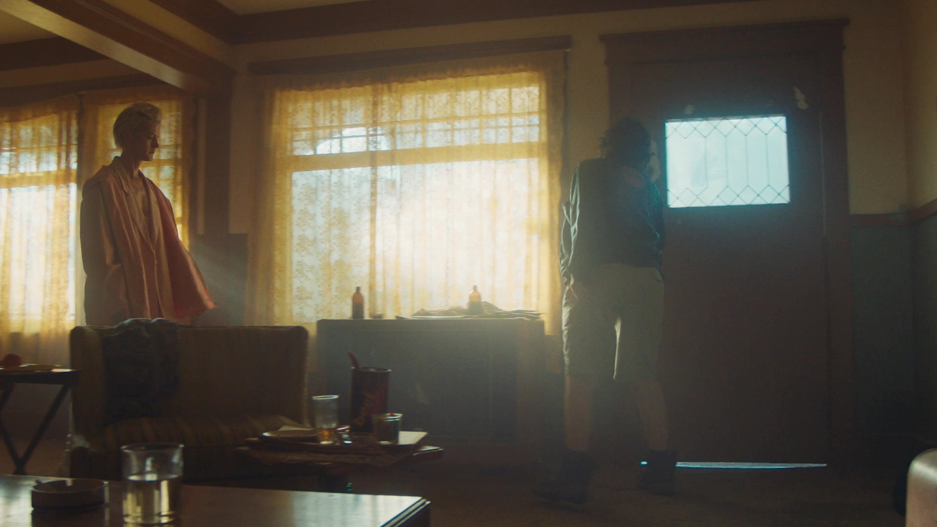 Two individuals engage in a somber conversation inside a dimly lit living room before one departs through the front door.