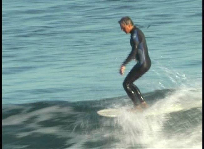 An older man in a wetsuit skillfully navigates ocean waves on a surfboard while also providing commentary on his experience. The footage alternates between close-up interview segments and wide shots of him surfing in a coastal environment.