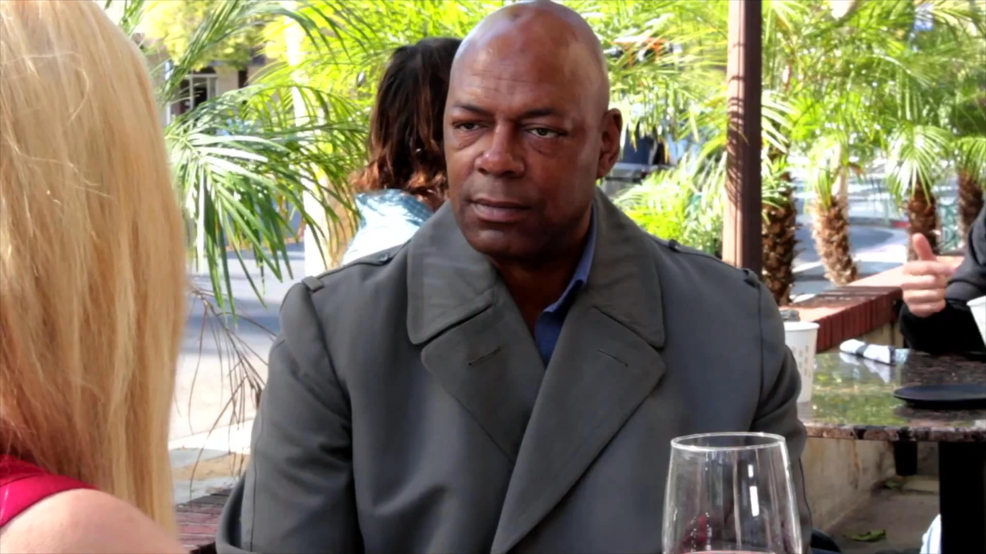 A woman and a man engage in a focused, seated conversation at an outdoor restaurant table. They maintain eye contact and exchange dialogue in a professional, casual dining setting.