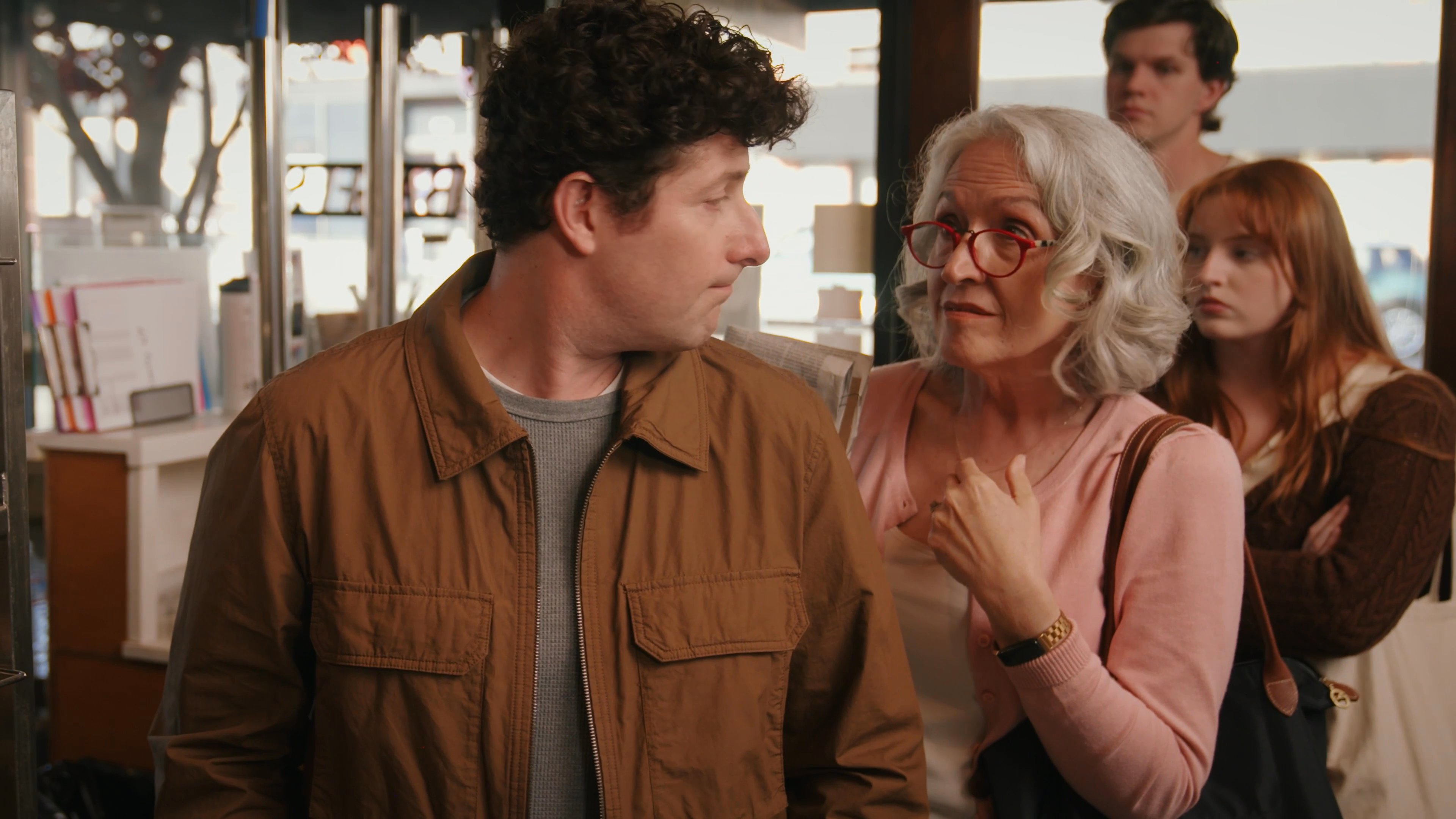 An older woman engages in a conversation with other customers inside a well-stocked bakery. The group interacts casually while browsing the shelves of fresh baked goods.