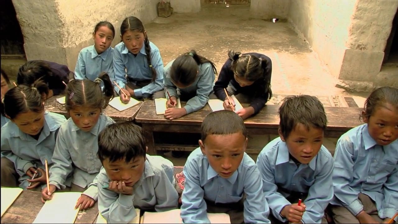 A teacher leads a group of young students in a rural classroom, demonstrating how to write on a chalkboard while the children practice their penmanship. The scene highlights the focus on literacy and education within a traditional, modest learning environment.