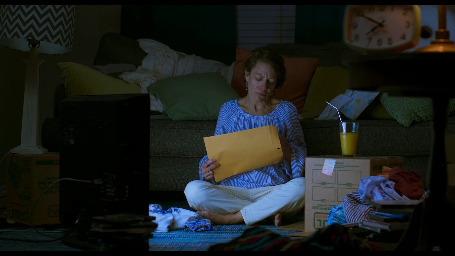 A woman sits on the floor of a dimly lit living room at night, opening and carefully reading the contents of a large manila envelope.