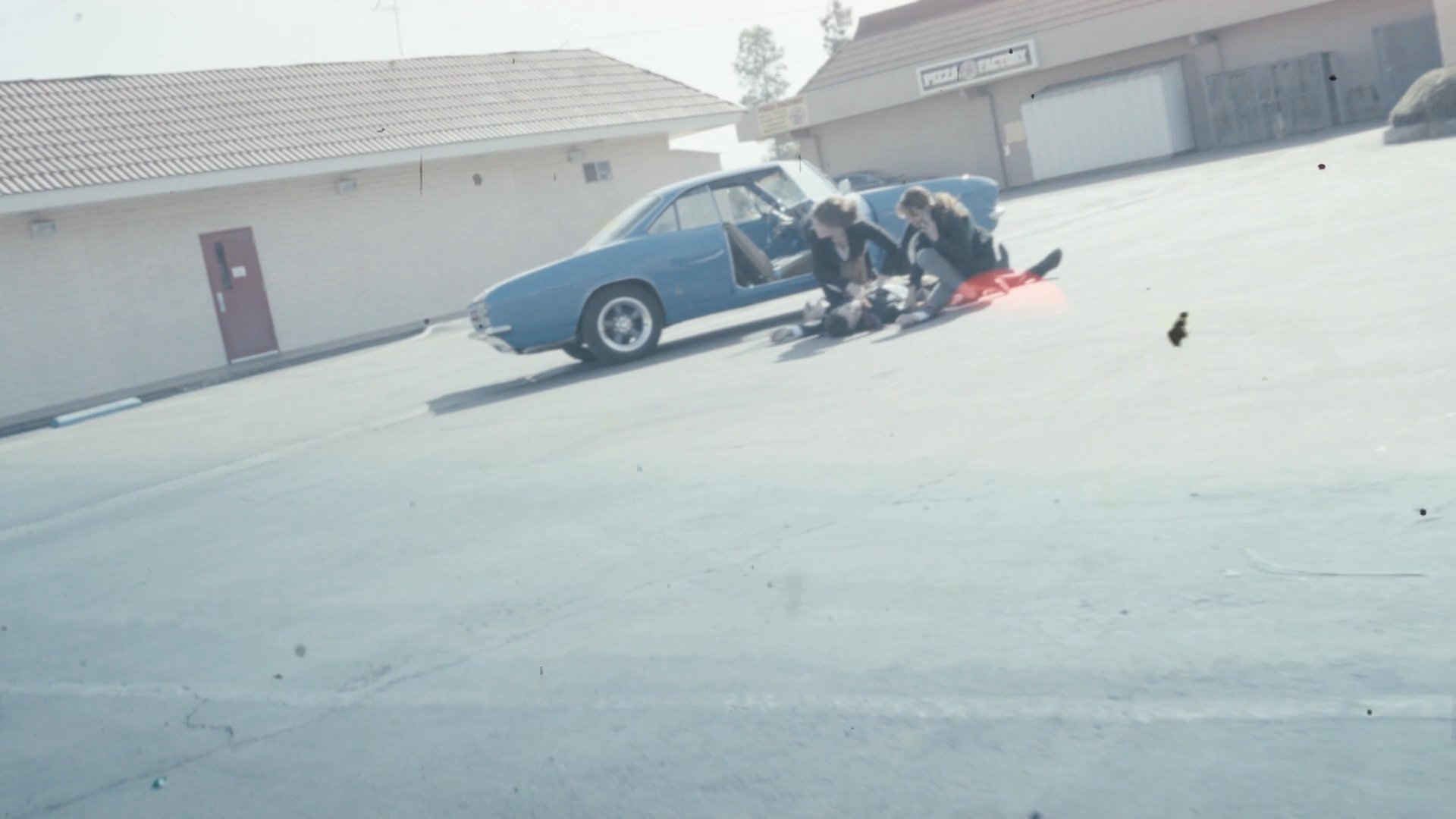 Two individuals attend to a person lying on the ground near a blue car in a parking lot, followed by a transition to a distant, hazy sunset over a landscape.