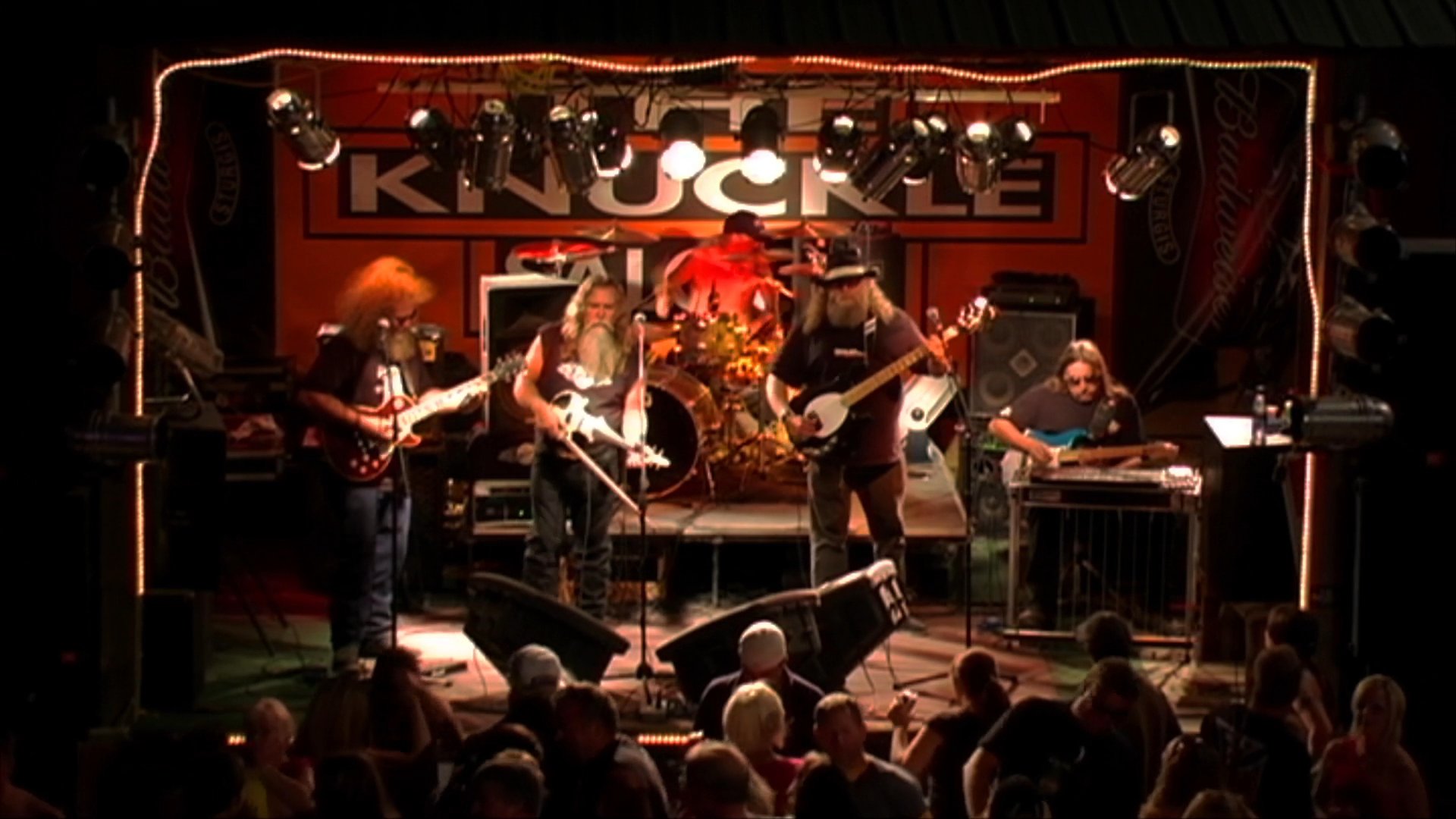 A live musical ensemble performs on a stage before an audience, featuring guitarists and a violinist in a dimly lit venue.