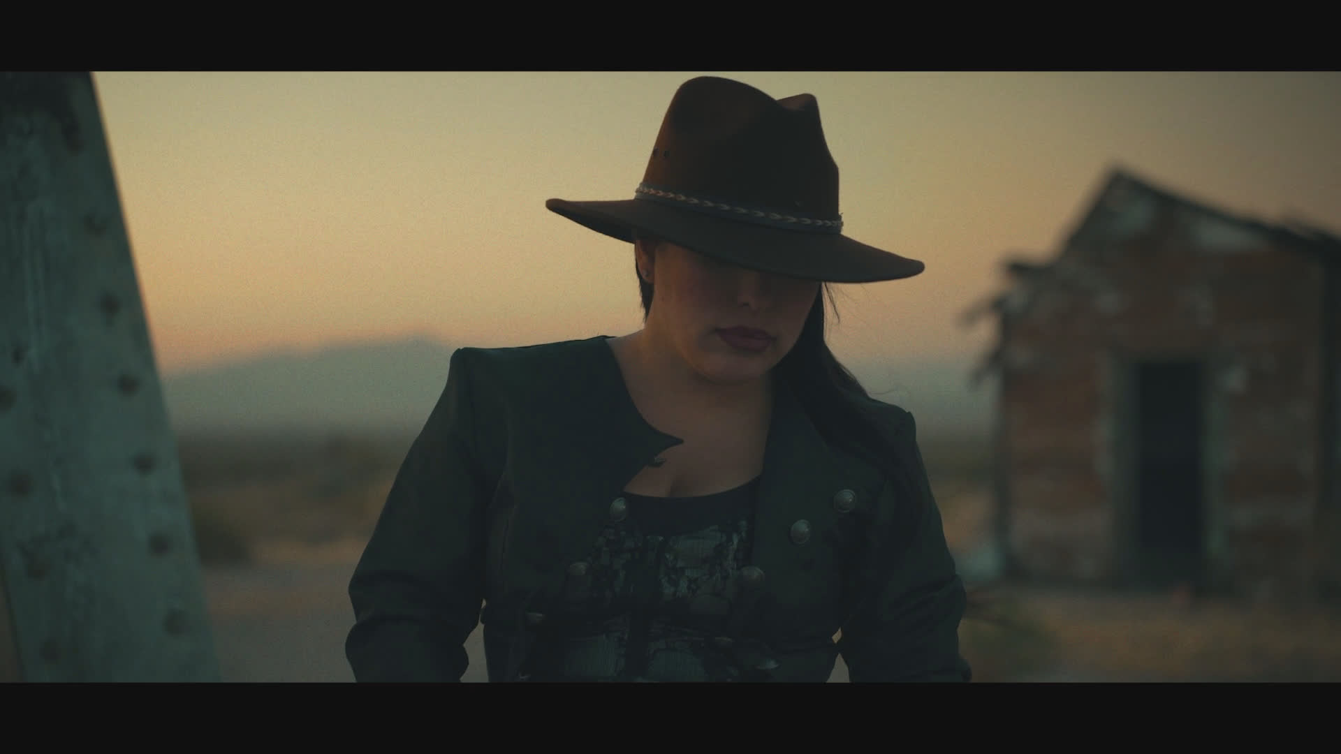 A woman in a black cowboy hat kneels beside a person lying on the ground at a remote outpost, then rises and speaks with grim resolve. The sunset-lit scene ends with her standing alone among weathered structures and open scrubland.