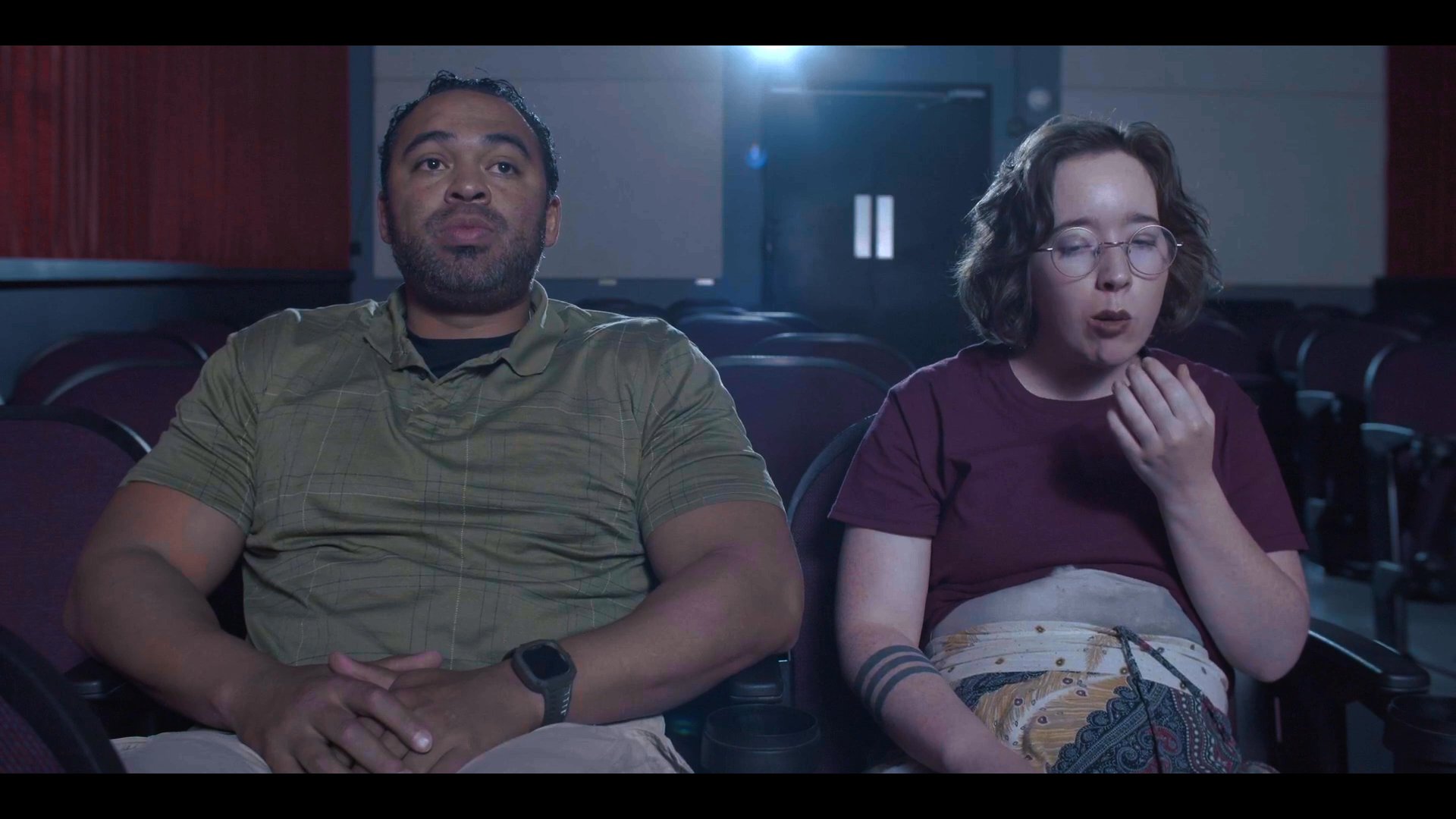 Patrons occupy a dark movie theater, snacking on popcorn while engaging in quiet conversation between film segments.