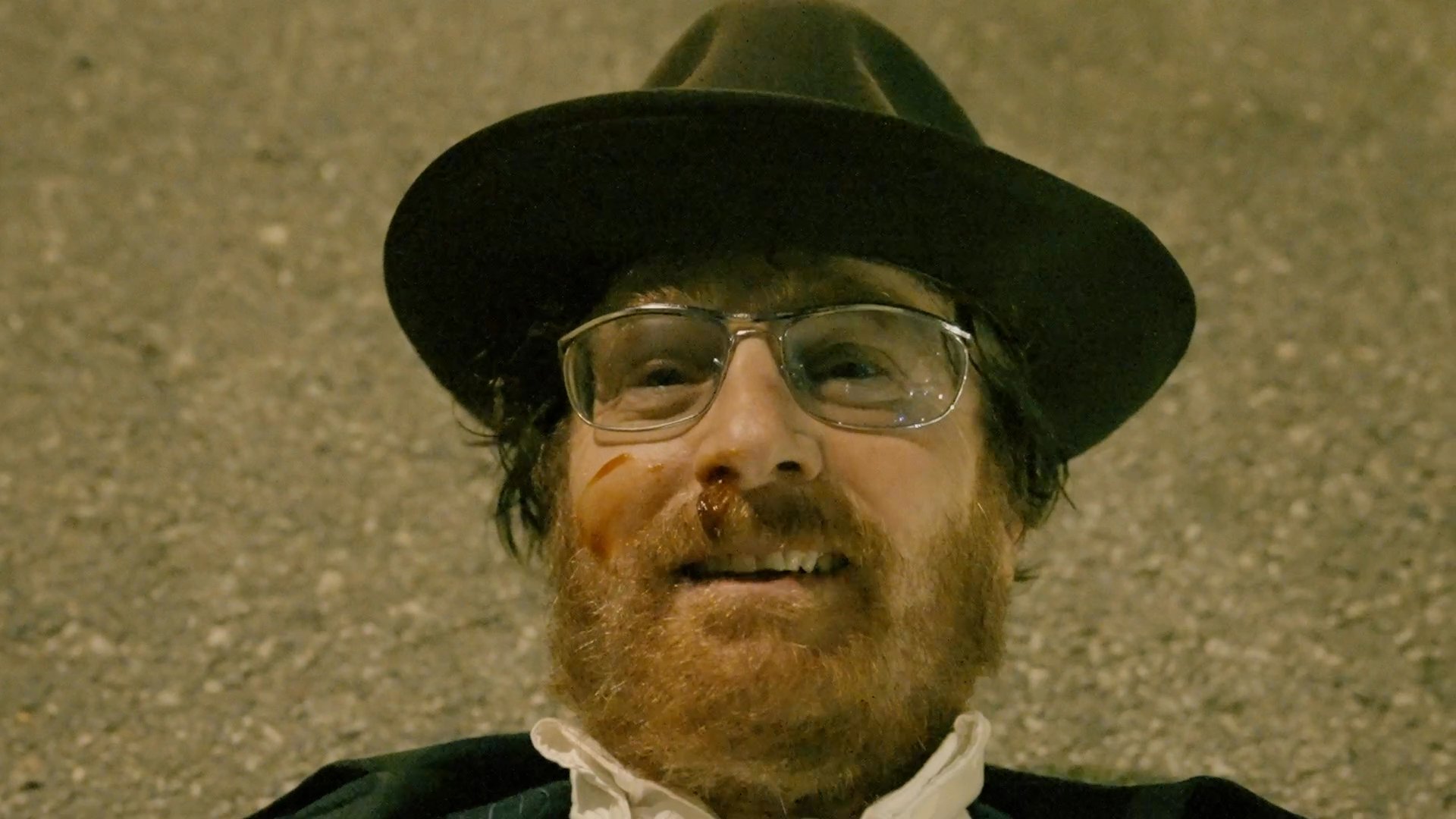 Two men exchange intense dialogue while sitting inside a parked car at night, interspersed with cuts to a man lying on the ground outdoors.