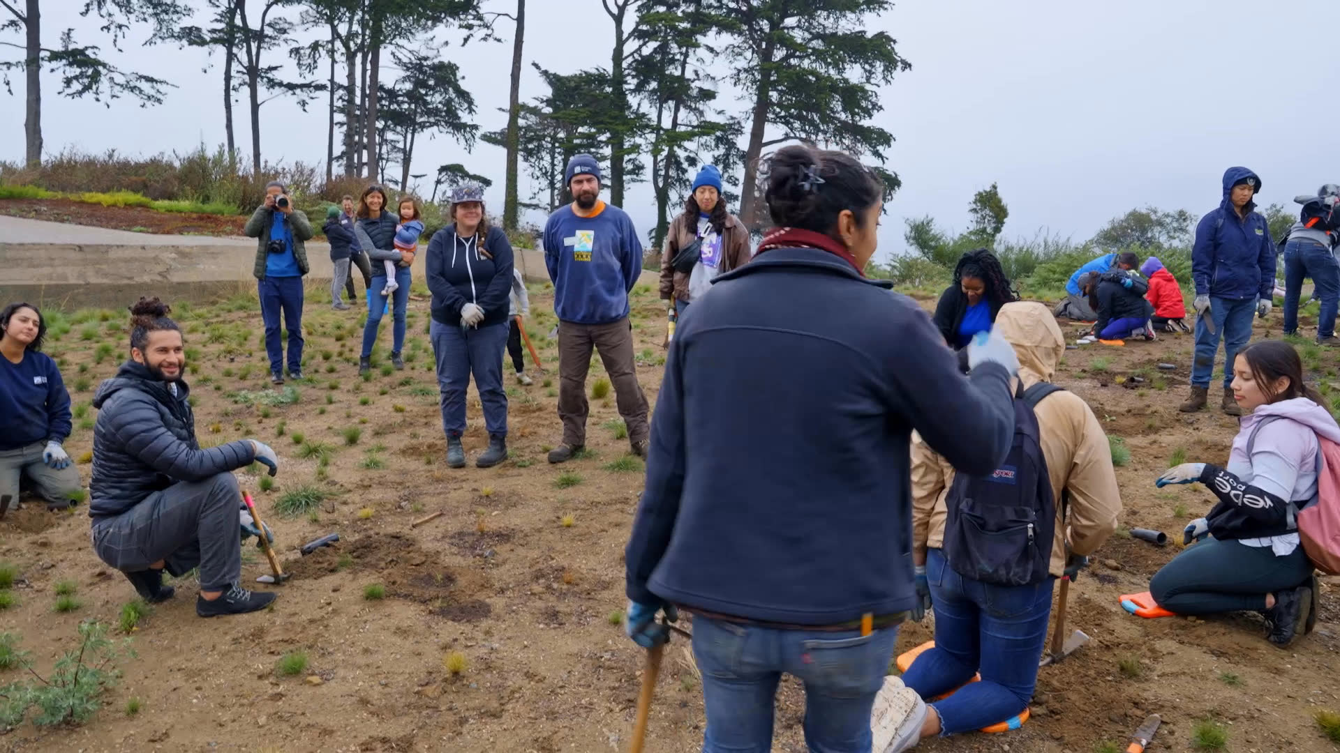 Volunteers dig and plant flowering greenery along a riverside, working together with hand tools as a group gathers nearby. A woman discusses the plants and the value of quiet observation, then speaks directly to camera.