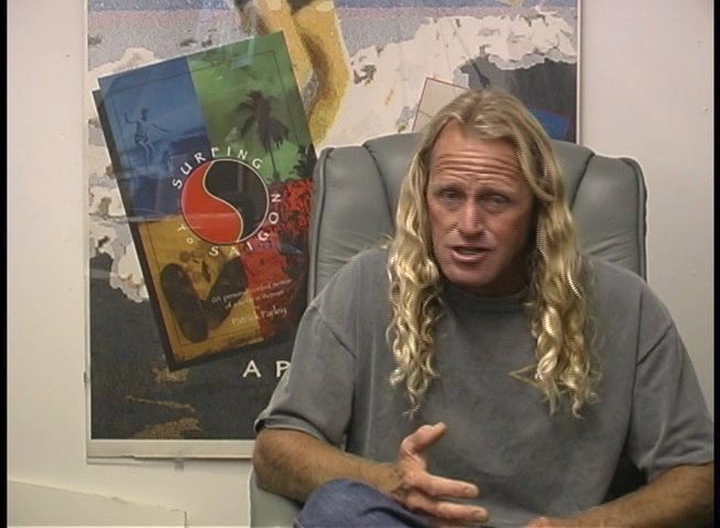 A long-haired man discusses professional filmmaking and camera equipment while seated before a surfing-themed backdrop, interspersed with shots of a professional video camera on a tripod by the coast.